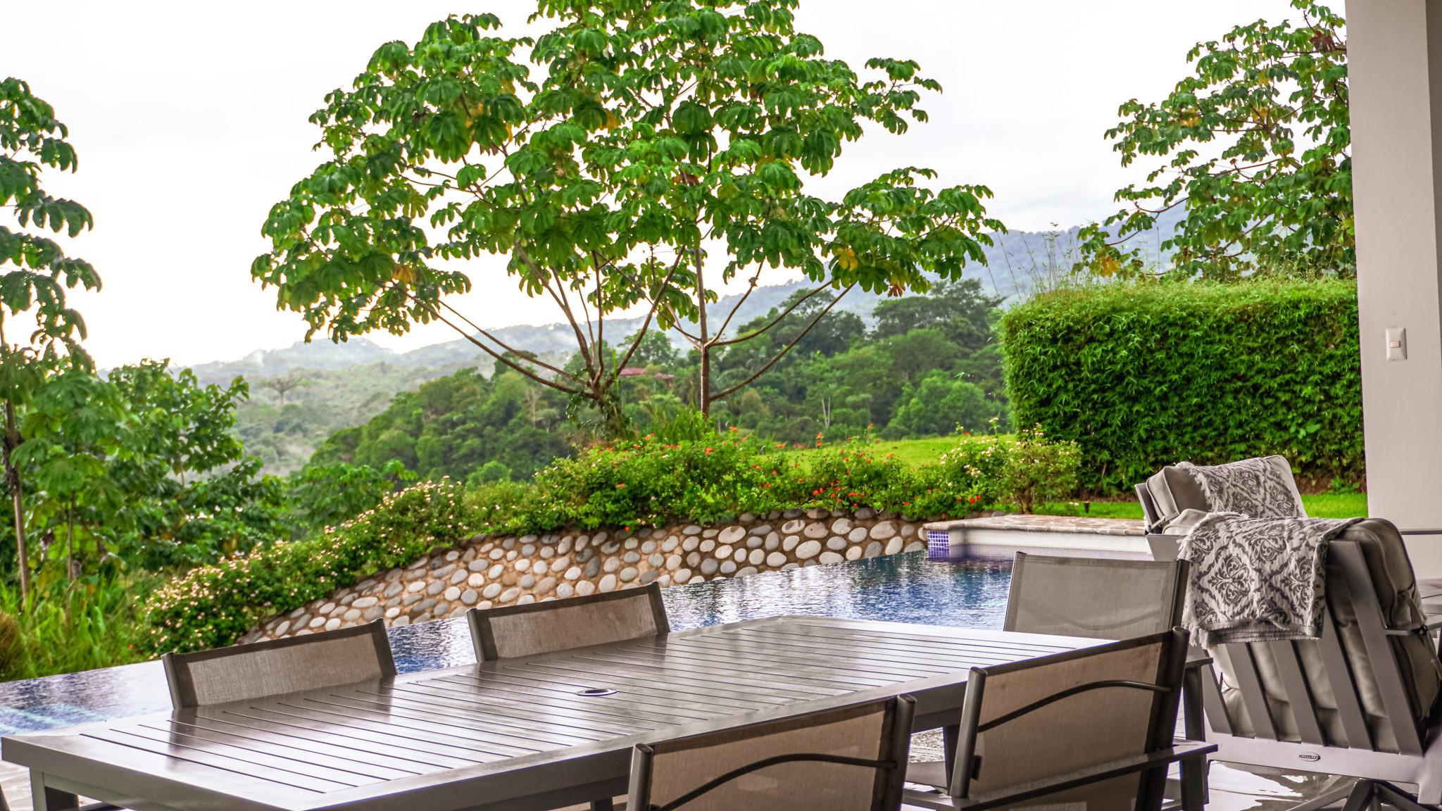 A covered outdoor dining area next to an infinity pool frames views of dense jungle and rolling mountains, bathed in natural light.