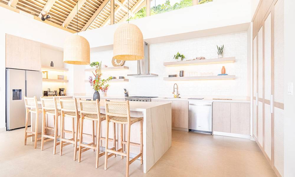 A bright, open-plan kitchen features a vaulted beamed ceiling, a large island with bar seating, light wood cabinetry, and a high transom window offering jungle views.