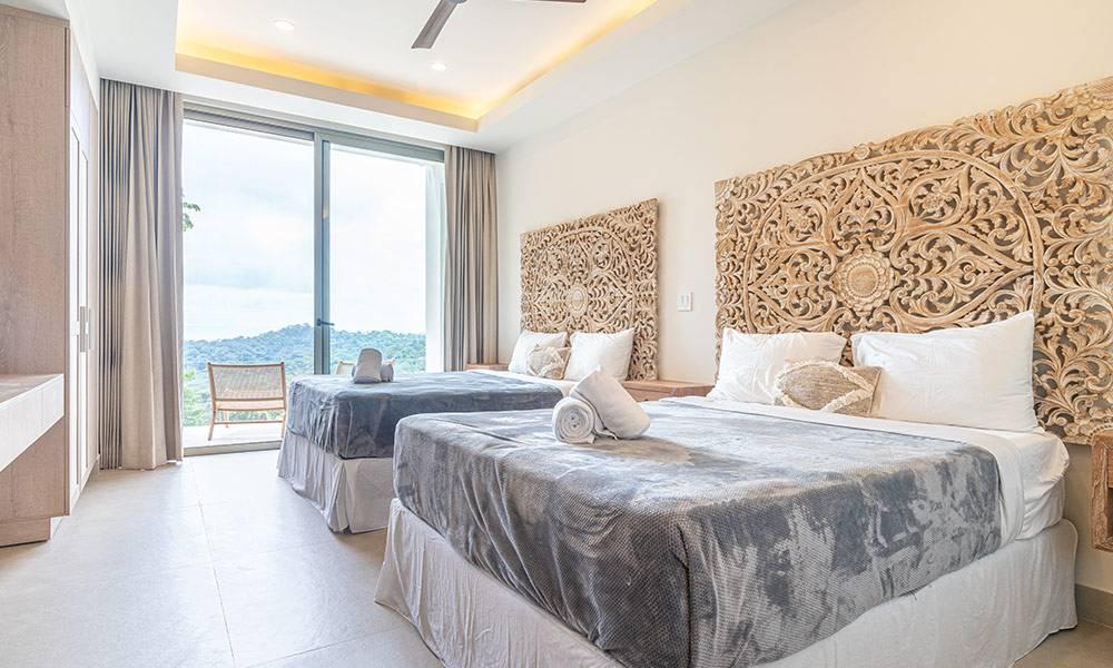 This bright bedroom features two queen beds with intricately carved headboards, receiving ample natural light from large sliding doors that open to a jungle and distant ocean view.