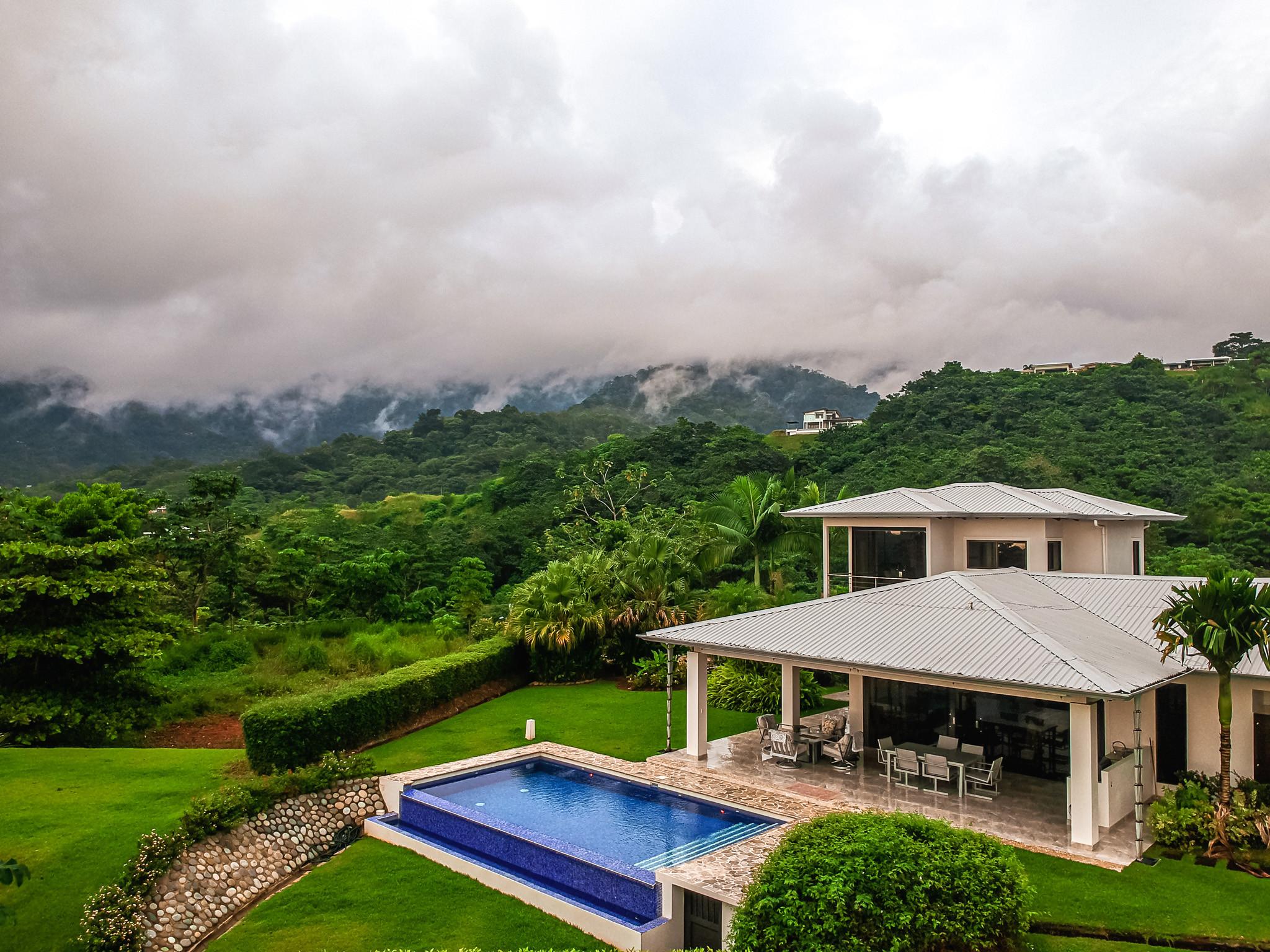 A modern white villa features a blue tiled pool and covered outdoor dining area, all set against a backdrop of lush, mist-covered jungle mountains.