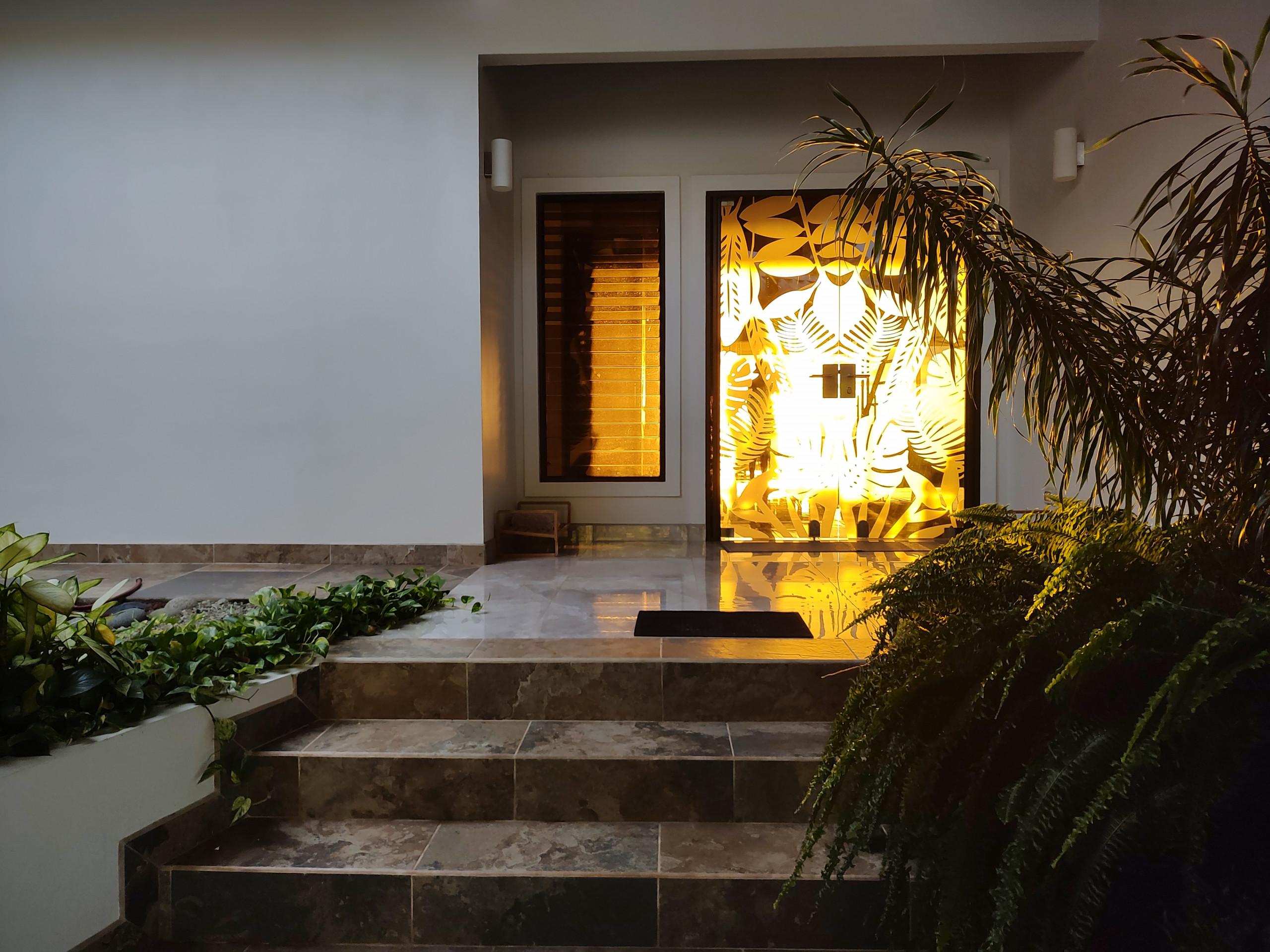The villa's main entrance features a tropical leaf-patterned glass door glowing with warm interior light, flanked by a louvered window, and accessed by tiled steps surrounded by lush exterior plants.