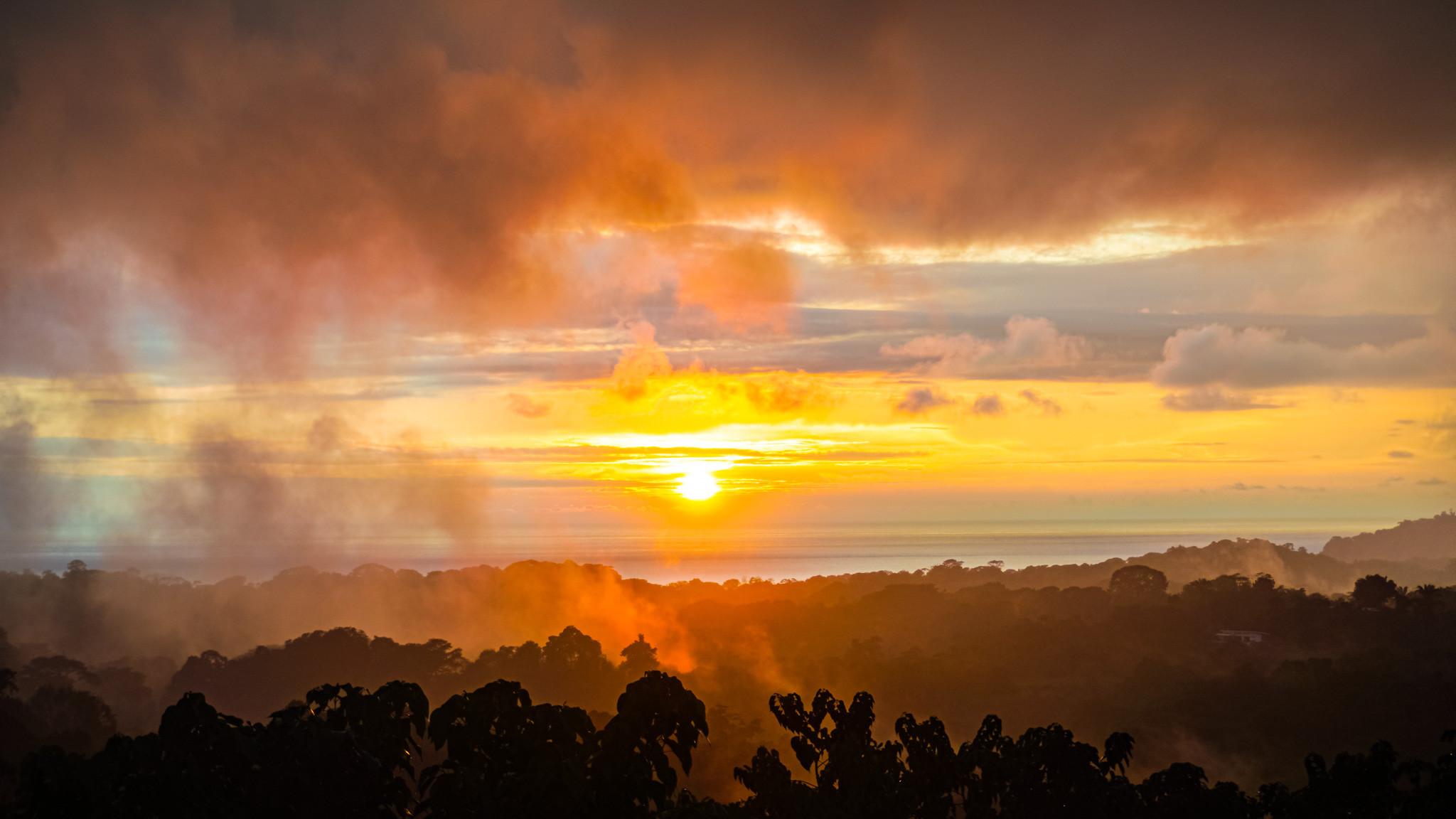 A vibrant sunset over the Pacific Ocean illuminates misty jungle-covered mountains.