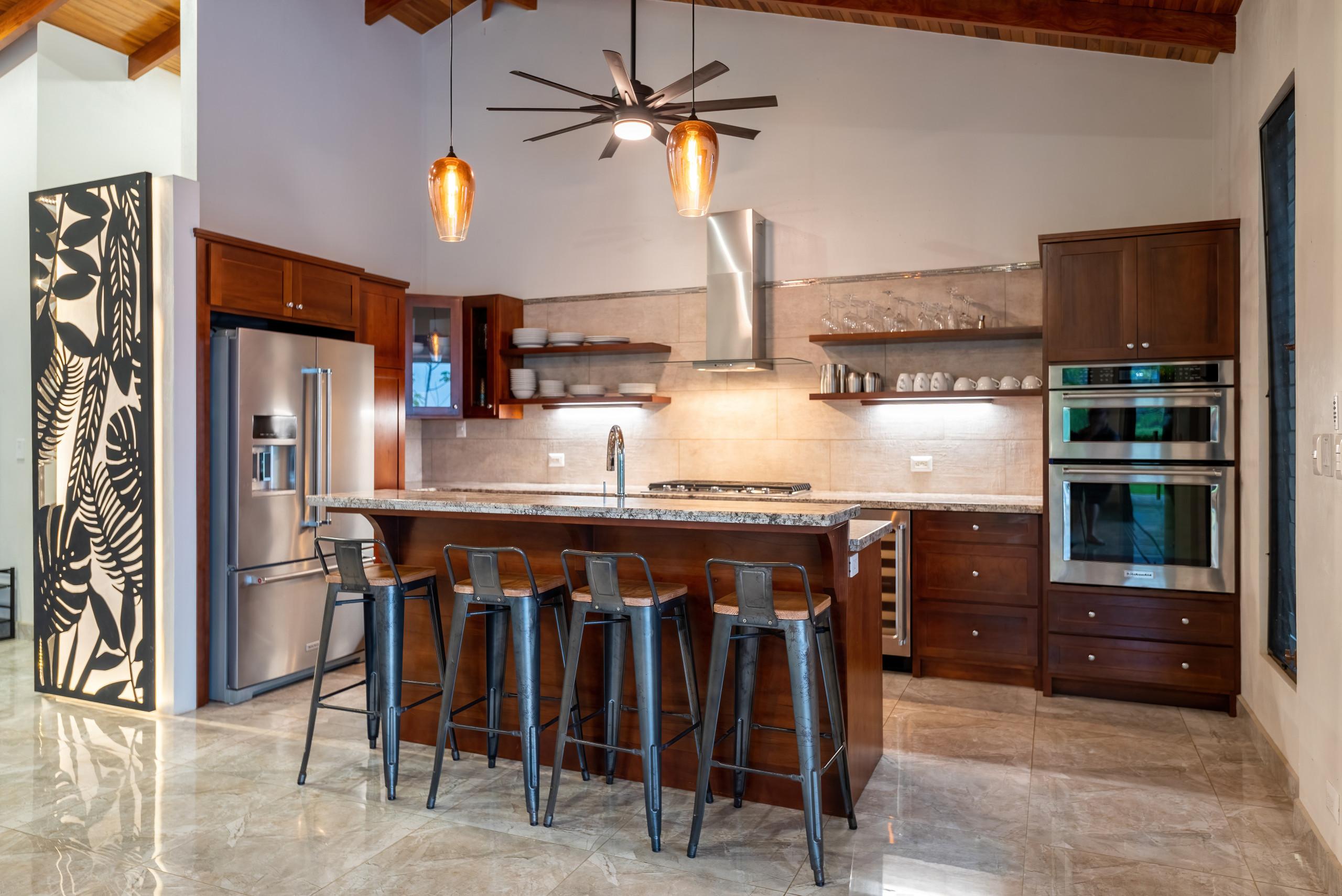 This open-concept kitchen features warm wood cabinetry, modern stainless steel appliances, and a granite island with seating under a vaulted ceiling, designed for cooking and socializing while connecting to the villa's outdoor environment.
