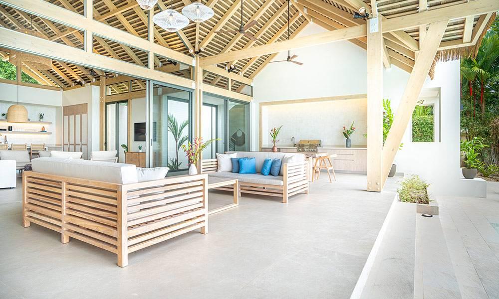 The expansive outdoor living space beneath a vaulted wooden ceiling features comfortable seating, an outdoor kitchen, and large glass doors that open to integrate with the surrounding jungle.