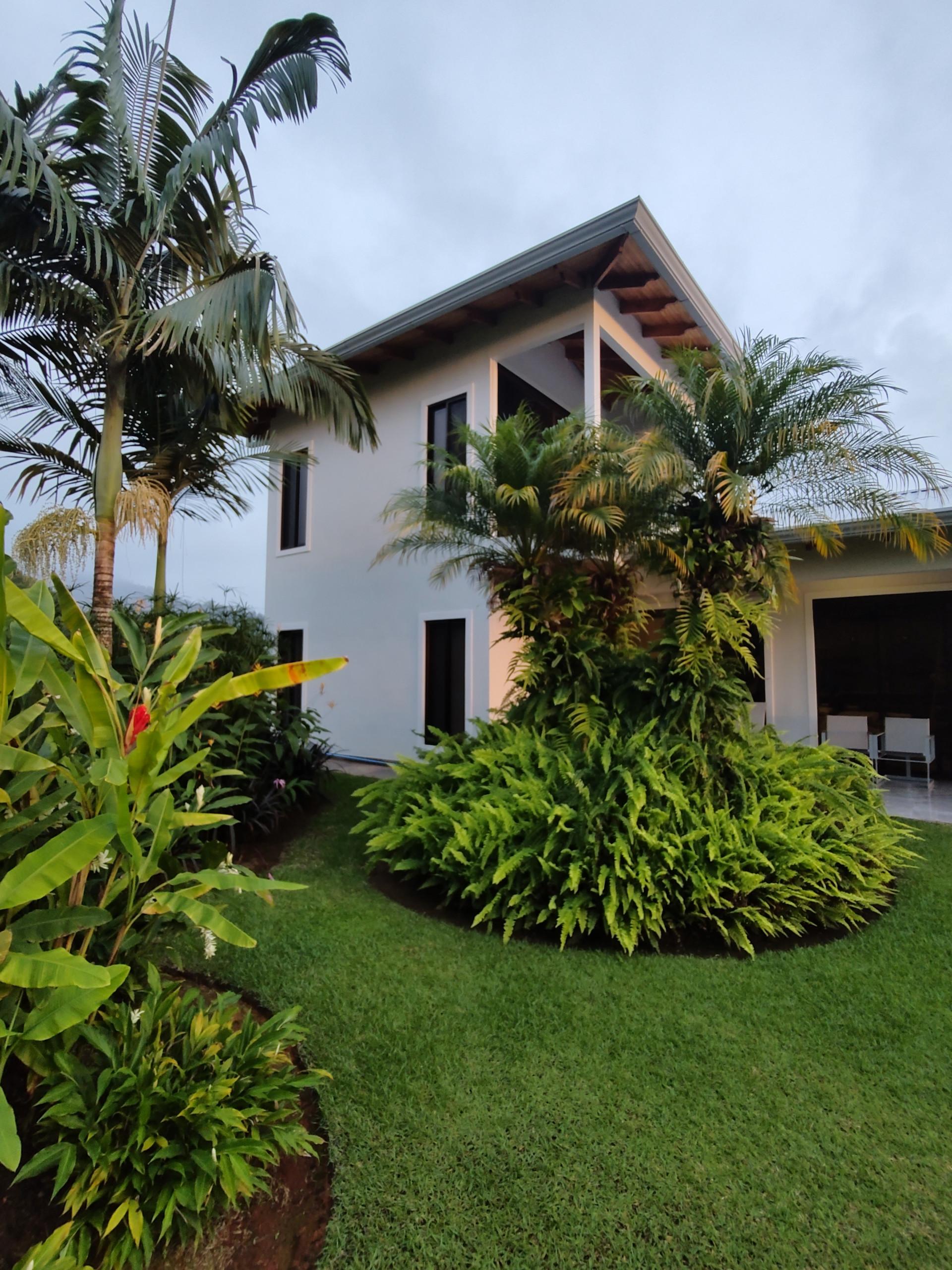 A two-story white villa with dark-framed windows and exposed wooden eaves, featuring an upper-level covered balcony and a ground-level covered parking area, surrounded by vibrant tropical landscaping and tall palm trees.