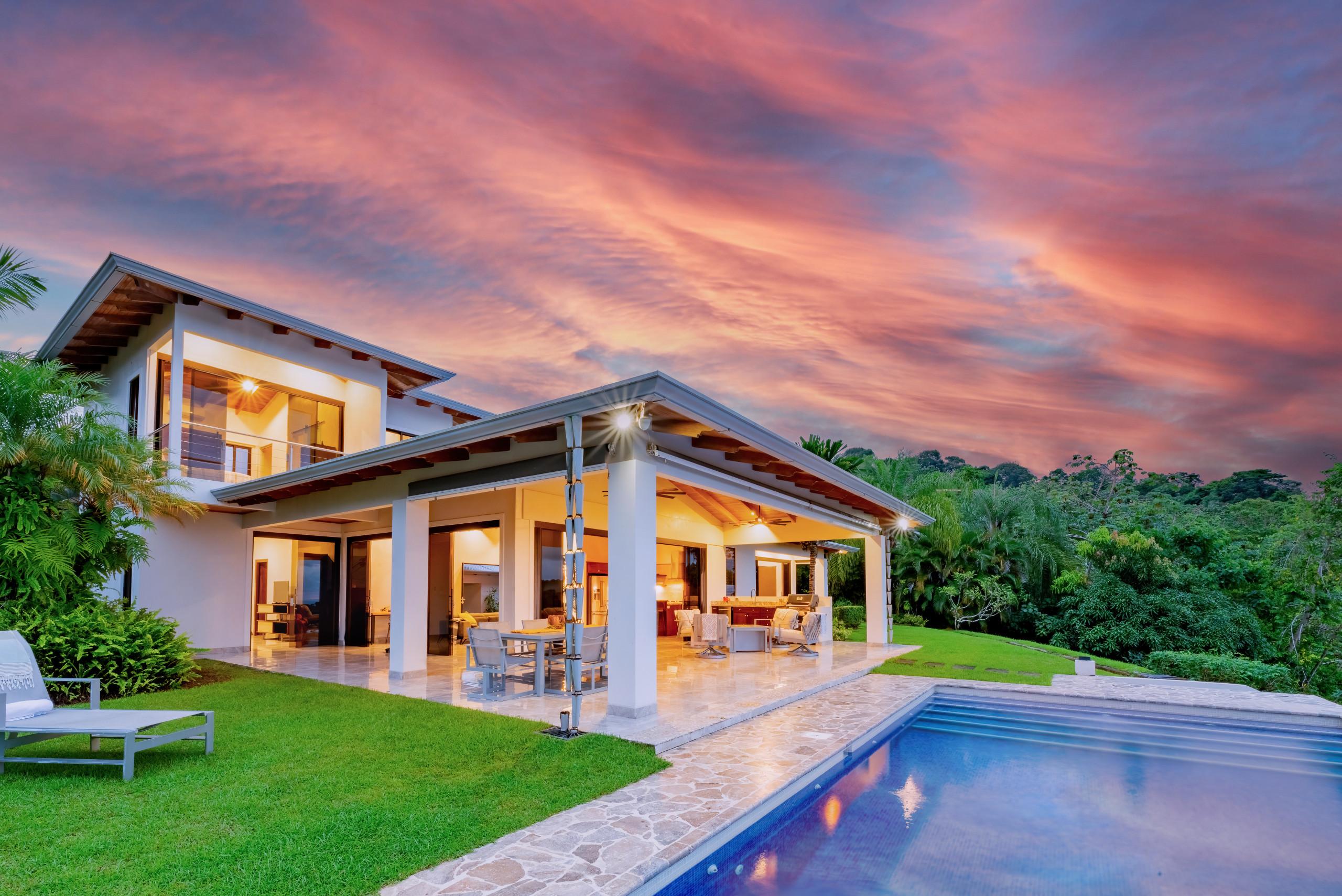 The contemporary two-story villa features an open-concept ground floor with glass walls, connecting to a covered outdoor living area and an infinity pool, all set against a backdrop of lush jungle and a vibrant sunset.