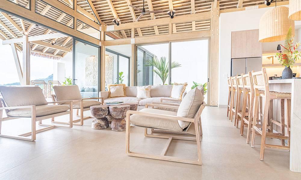 This expansive open-plan living area features high vaulted wooden ceilings and a seamless flow to an integrated kitchen bar, all illuminated by wide glass doors offering connection to the outdoor landscape.