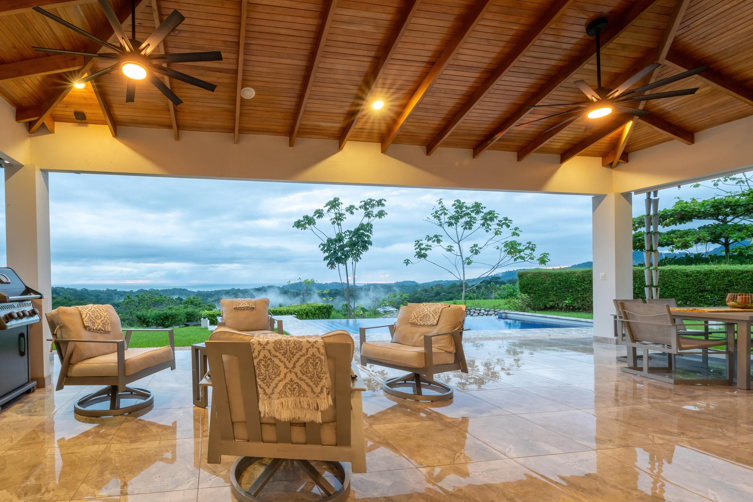 The covered outdoor terrace features a high wood-paneled ceiling, comfortable seating, and an outdoor dining area, all opening to an infinity pool with expansive jungle and ocean views.