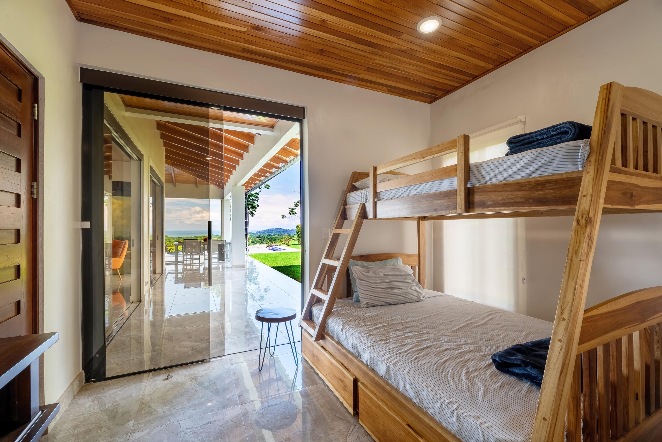 This bunk room features a custom wooden bunk bed and a slatted wood ceiling, with a large glass door opening to a terrace offering natural light and views of the pool, jungle, and ocean.