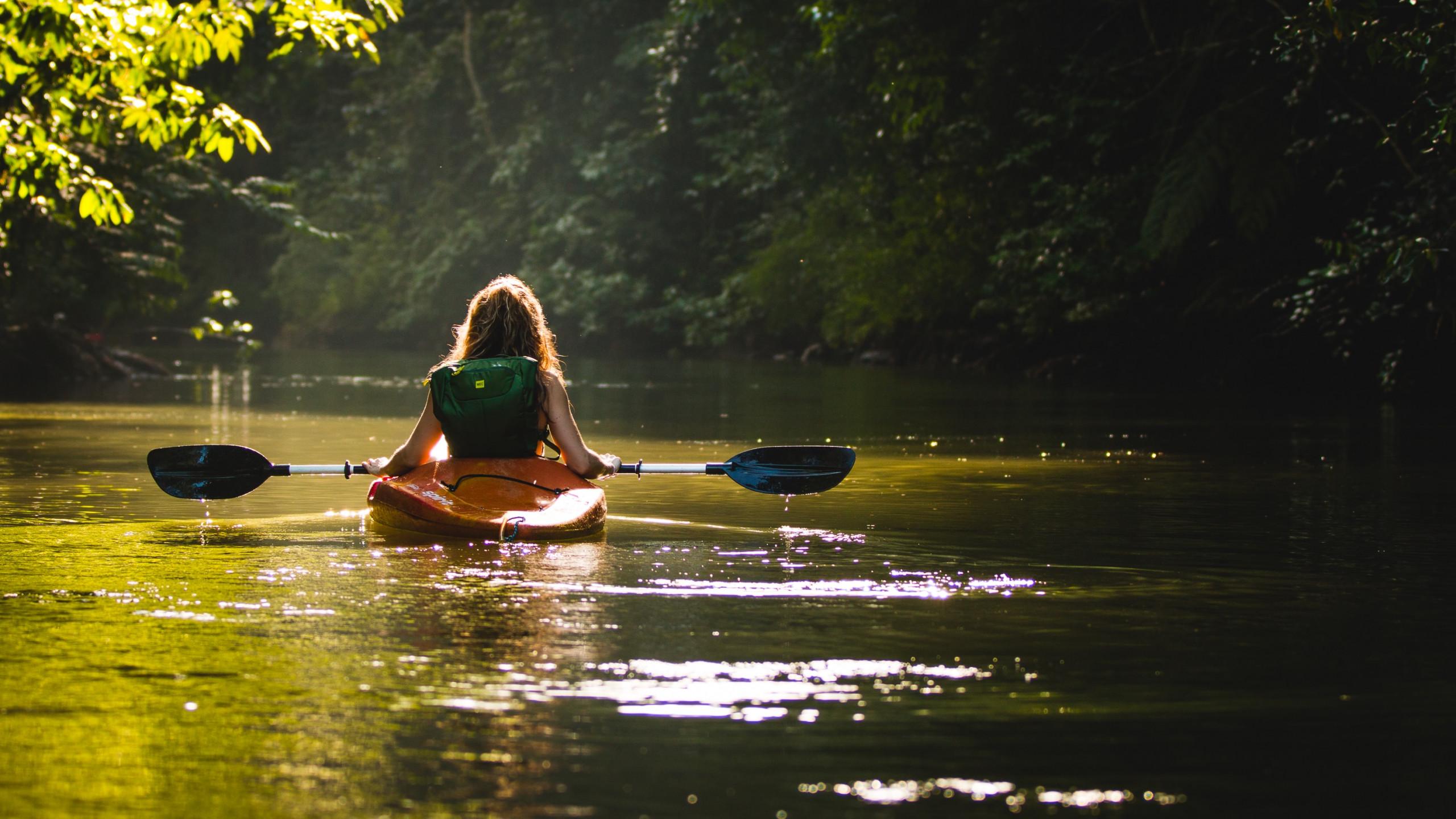 A person in a kayak paddles on a tranquil, sunlit river bordered by dense tropical foliage.