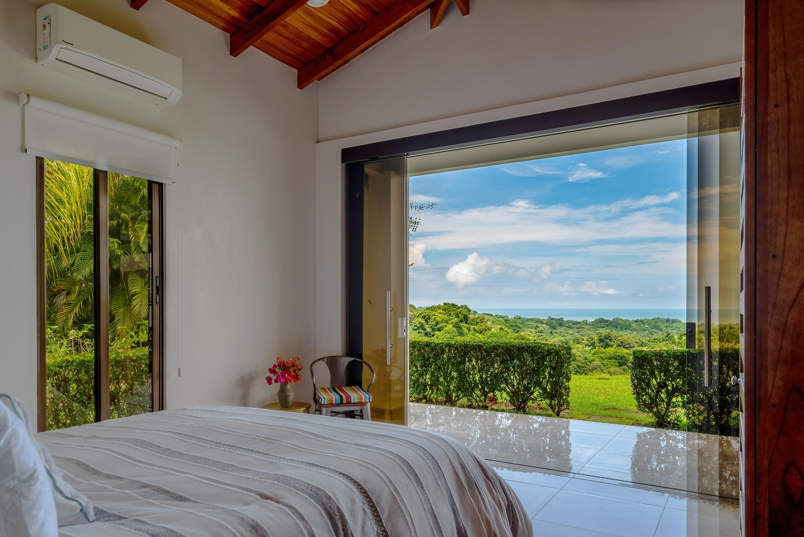A bright bedroom with a queen-size bed and vaulted wooden ceiling opens through wide glass doors to an outdoor area showcasing jungle and distant ocean views.