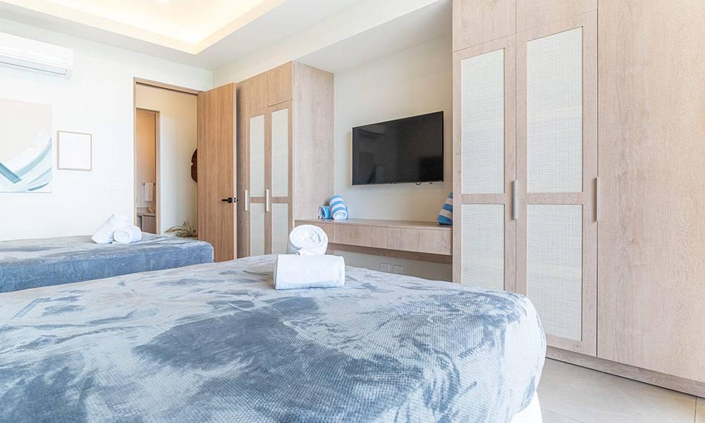 This bright guest room features two queen beds, built-in light wood cabinetry with textured panels, a wall-mounted television, and an adjacent private bathroom.