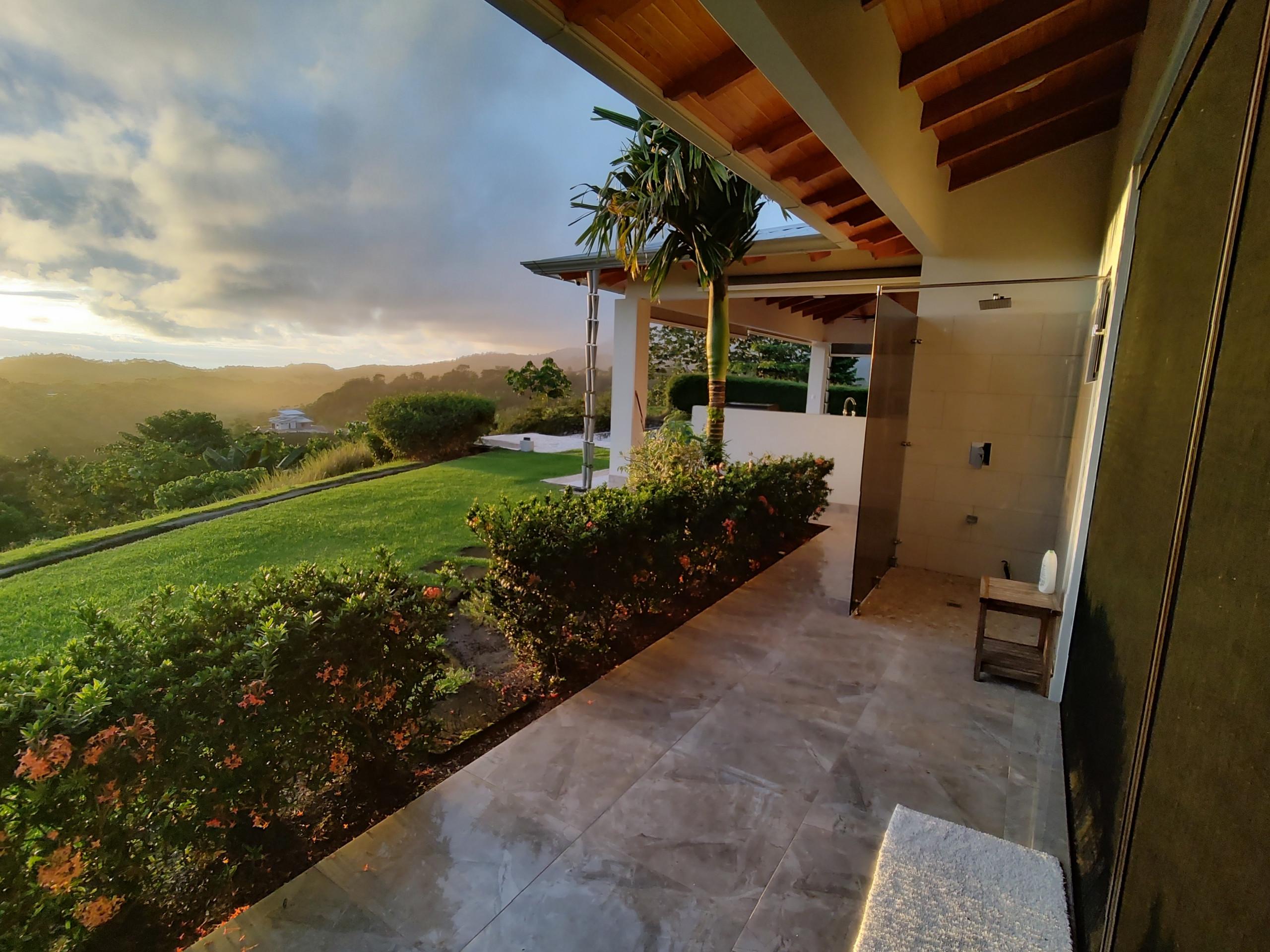 A modern tiled outdoor terrace with a rain shower head is covered by a wood-paneled ceiling, overlooking rolling, jungle-covered mountains illuminated by a sunset sky.