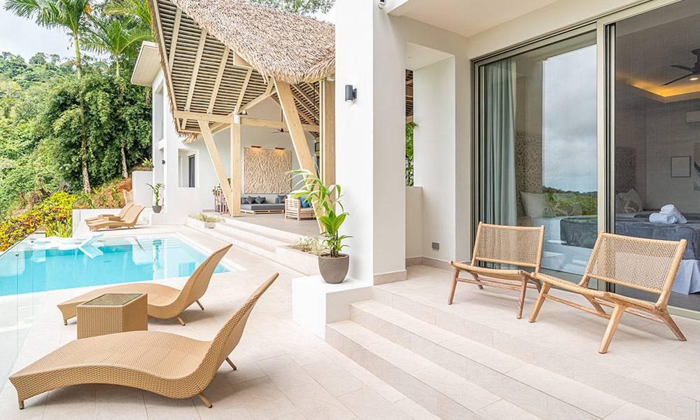 A bright outdoor terrace with an infinity pool and lounge chairs connects to a bedroom through sliding glass doors, while a thatched-roof open-air lounge overlooks the jungle-covered hills.