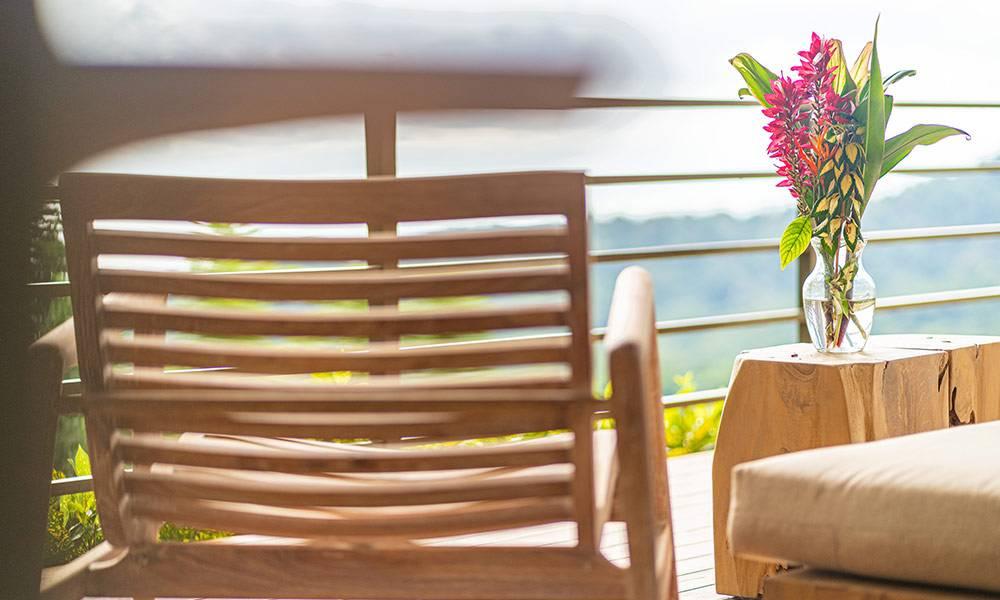 The outdoor terrace features wooden lounge furniture, vibrant tropical flowers, and expansive natural views.