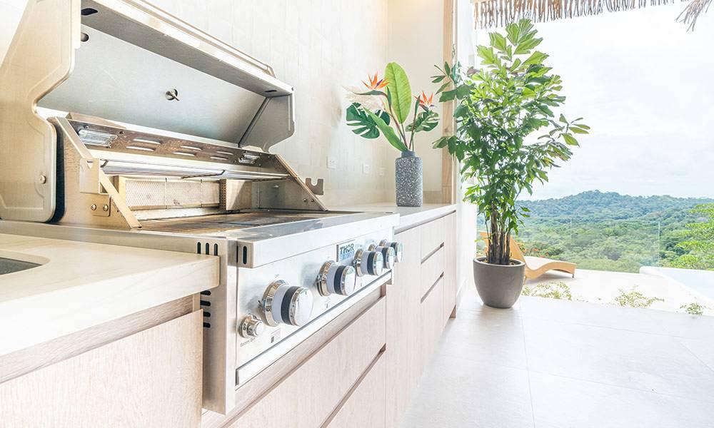A bright outdoor kitchen area, featuring a built-in stainless steel grill and light wood cabinetry, opens to extensive views of the tropical jungle.