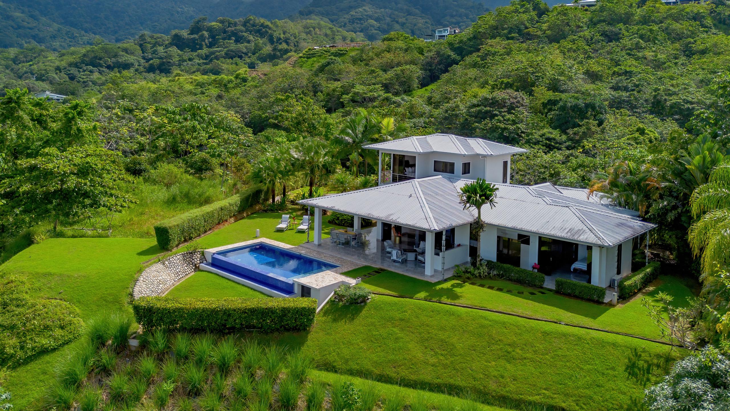 An aerial view shows the modern villa with a pool and covered porch, set on a lush green hillside overlooking dense jungle mountains.