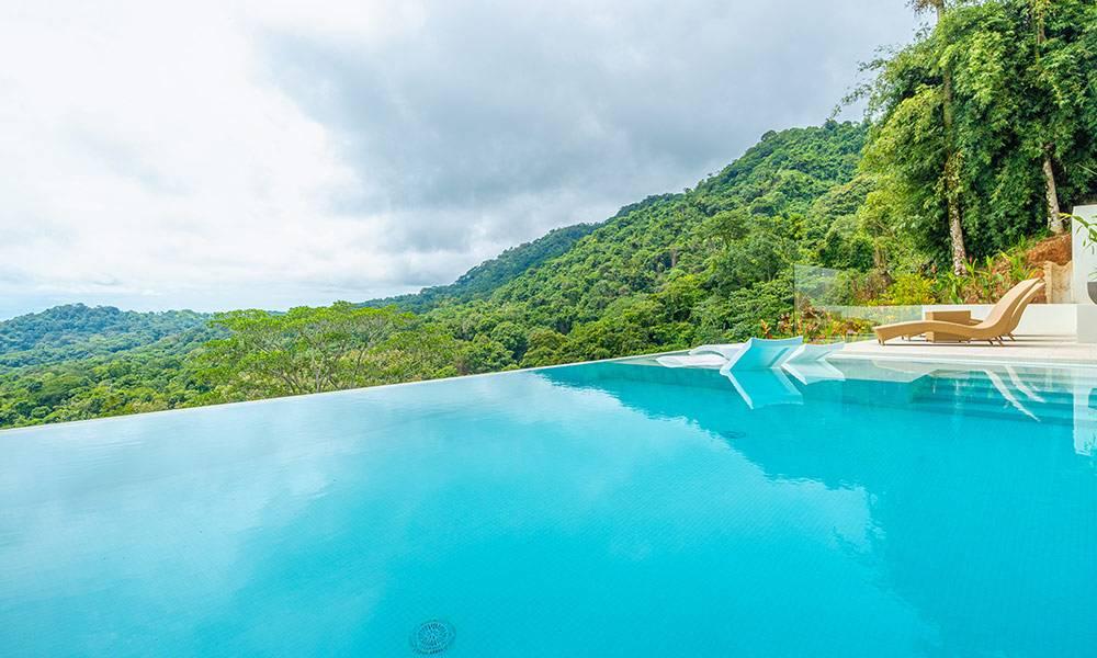 The infinity pool overlooks dense jungle hills stretching to the horizon, with a bright pool deck and chaise loungers.