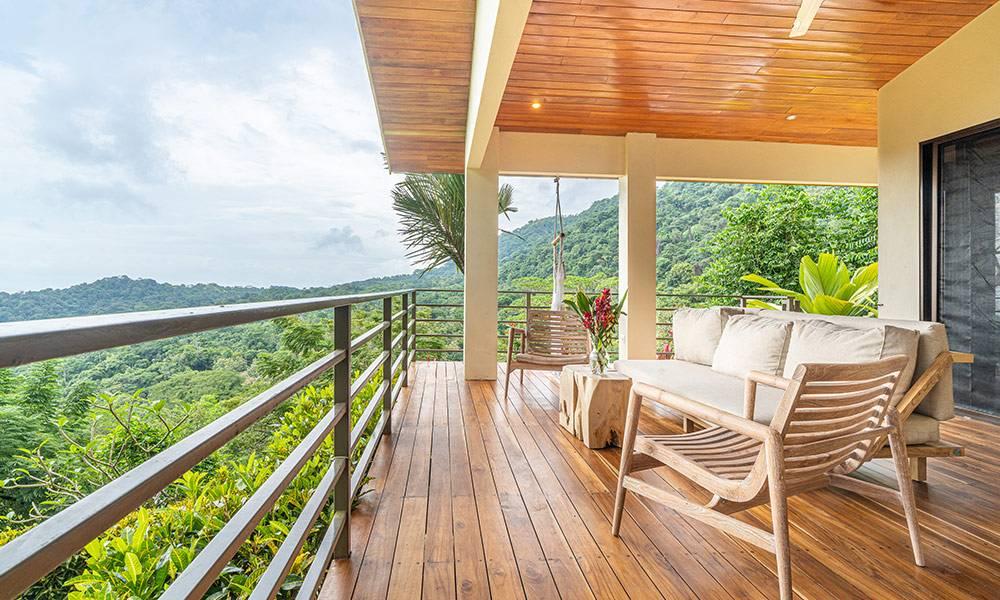 A covered wooden terrace with a plank floor and ceiling hosts casual seating, opening to extensive views of the jungle-covered hills under natural light.