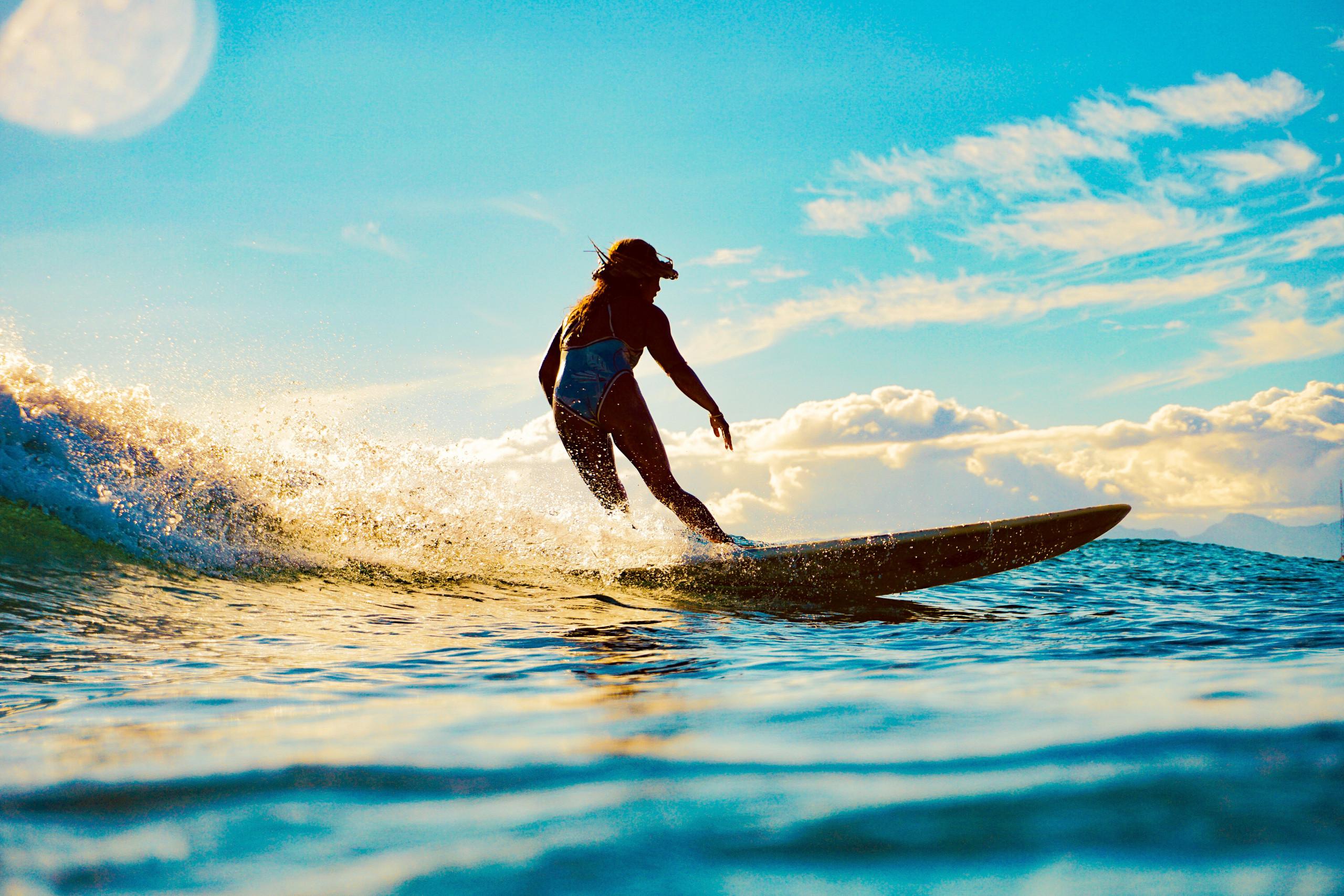 A person on a surfboard rides a breaking wave illuminated by golden light, with the ocean extending to distant mountains under a bright, cloudy sky.
