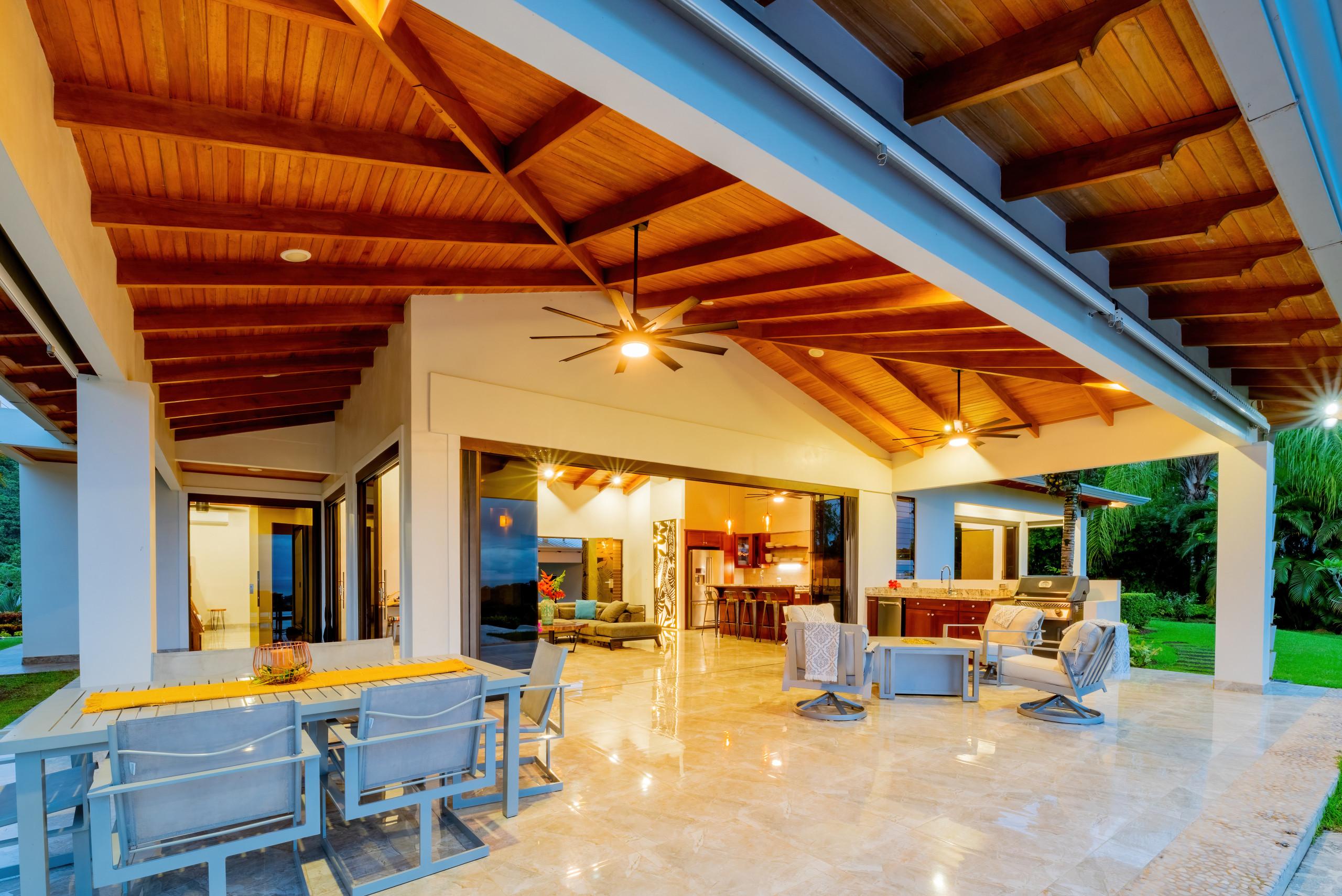 A covered outdoor terrace with a wooden beamed ceiling and tiled floor provides an outdoor dining area, a barbecue grill, and lounge seating, connecting to the villa's interior and surrounded by jungle.