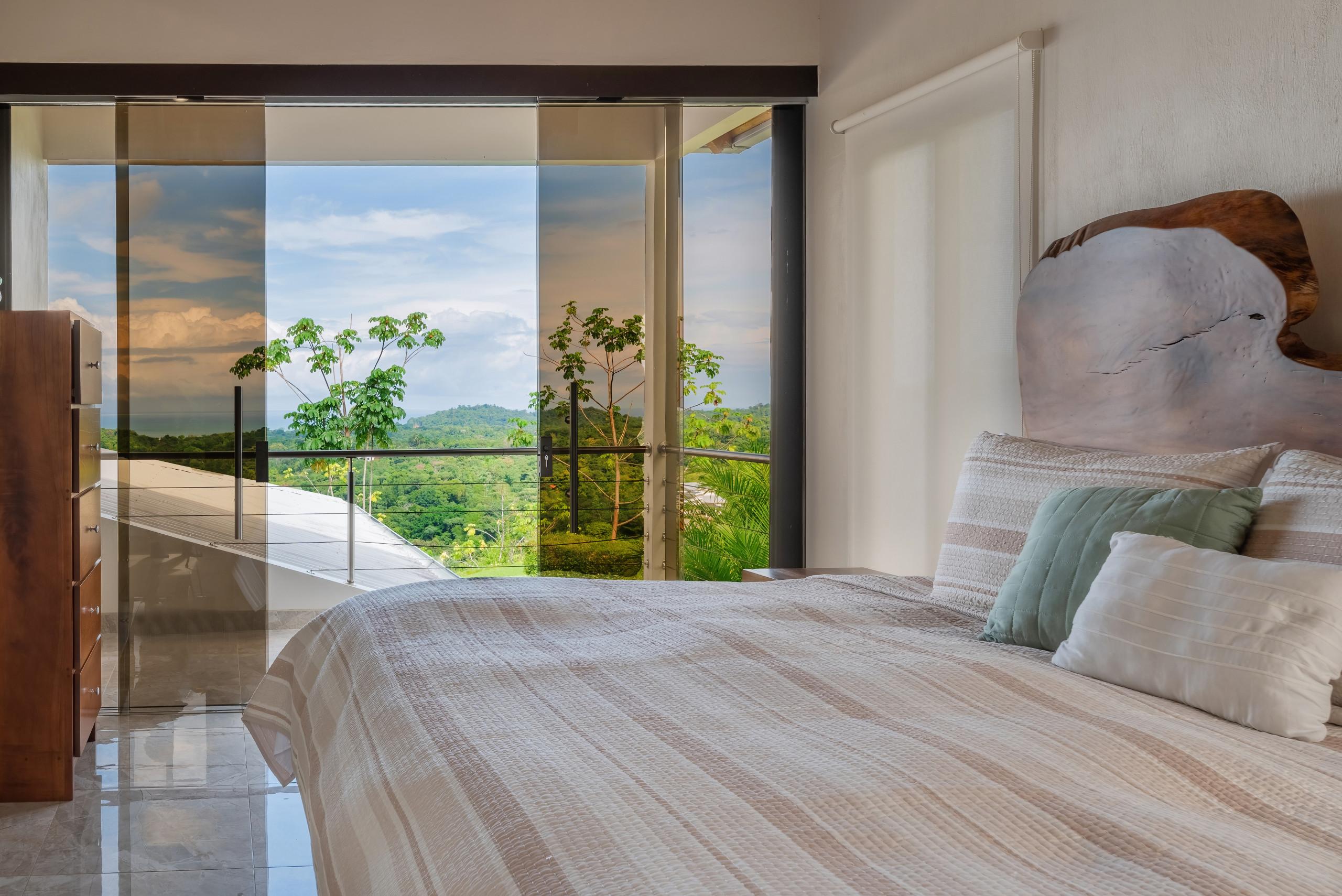 This bedroom features a natural wood headboard and receives abundant light through full-height glass doors that open to a balcony with jungle and distant ocean views.