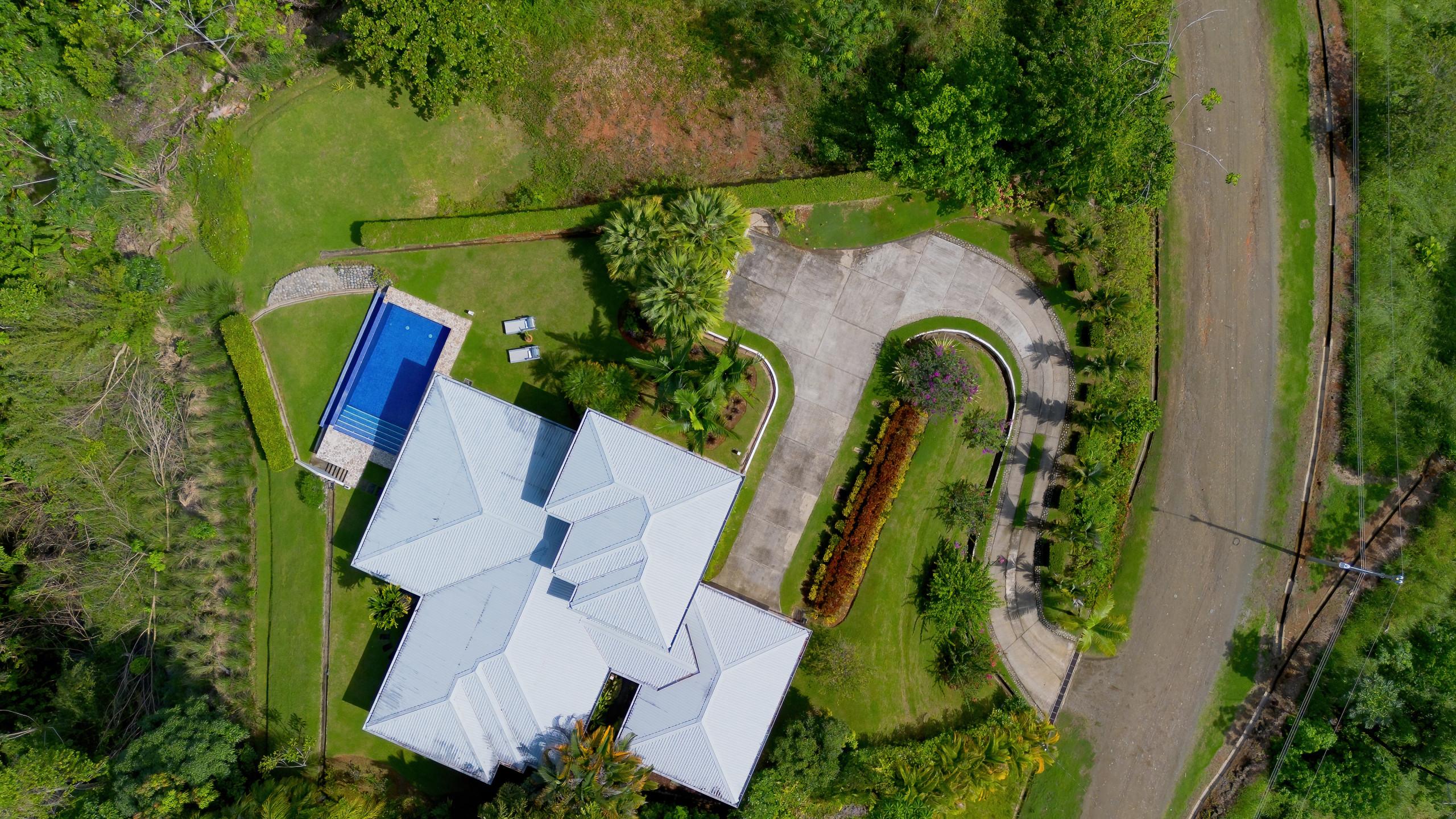 An aerial view reveals the villa's multi-roofline design, a bright blue swimming pool, and a winding paved driveway, all surrounded by lush green lawns and dense tropical jungle.