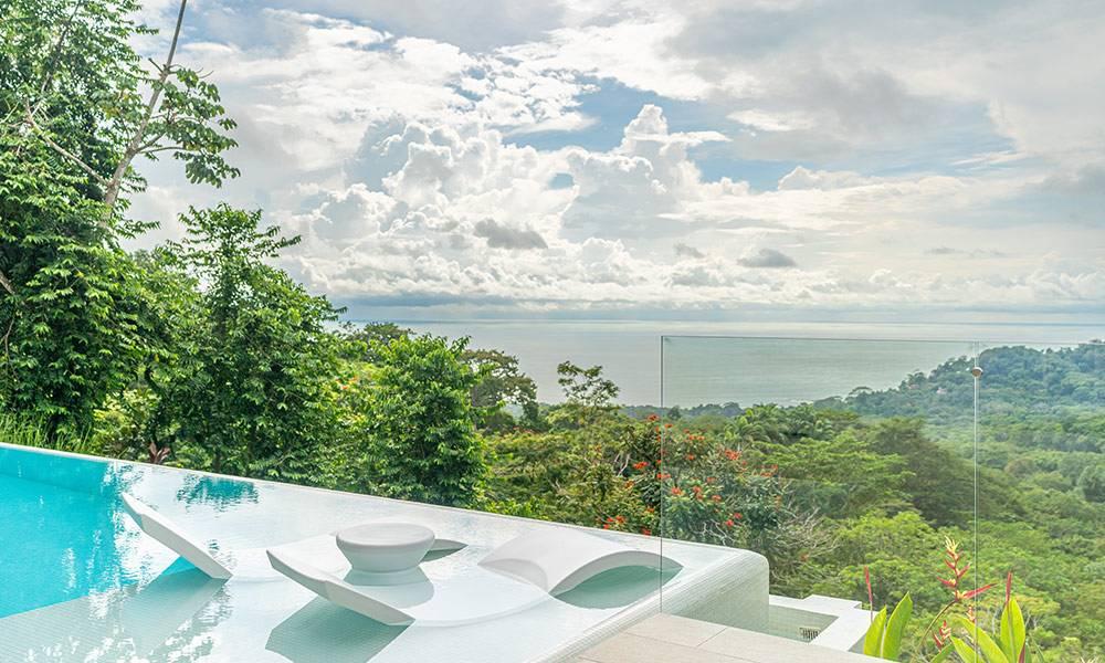The infinity pool features integrated submerged loungers and offers expansive views of the jungle and ocean under a bright, cloudy sky.