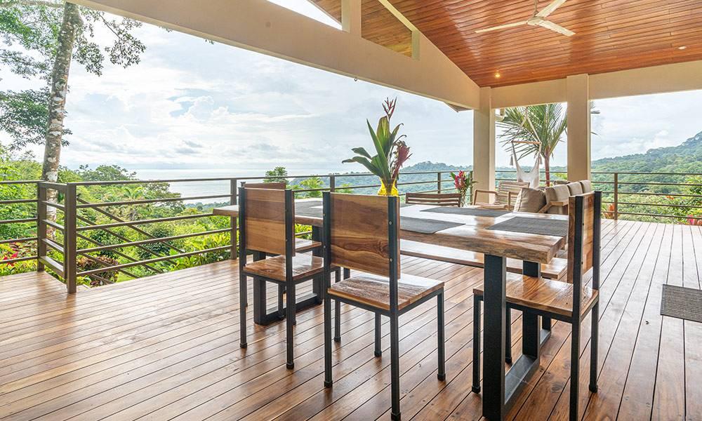 A spacious covered wooden deck features a natural wood dining table and seating area, offering expansive views of the jungle and distant ocean.