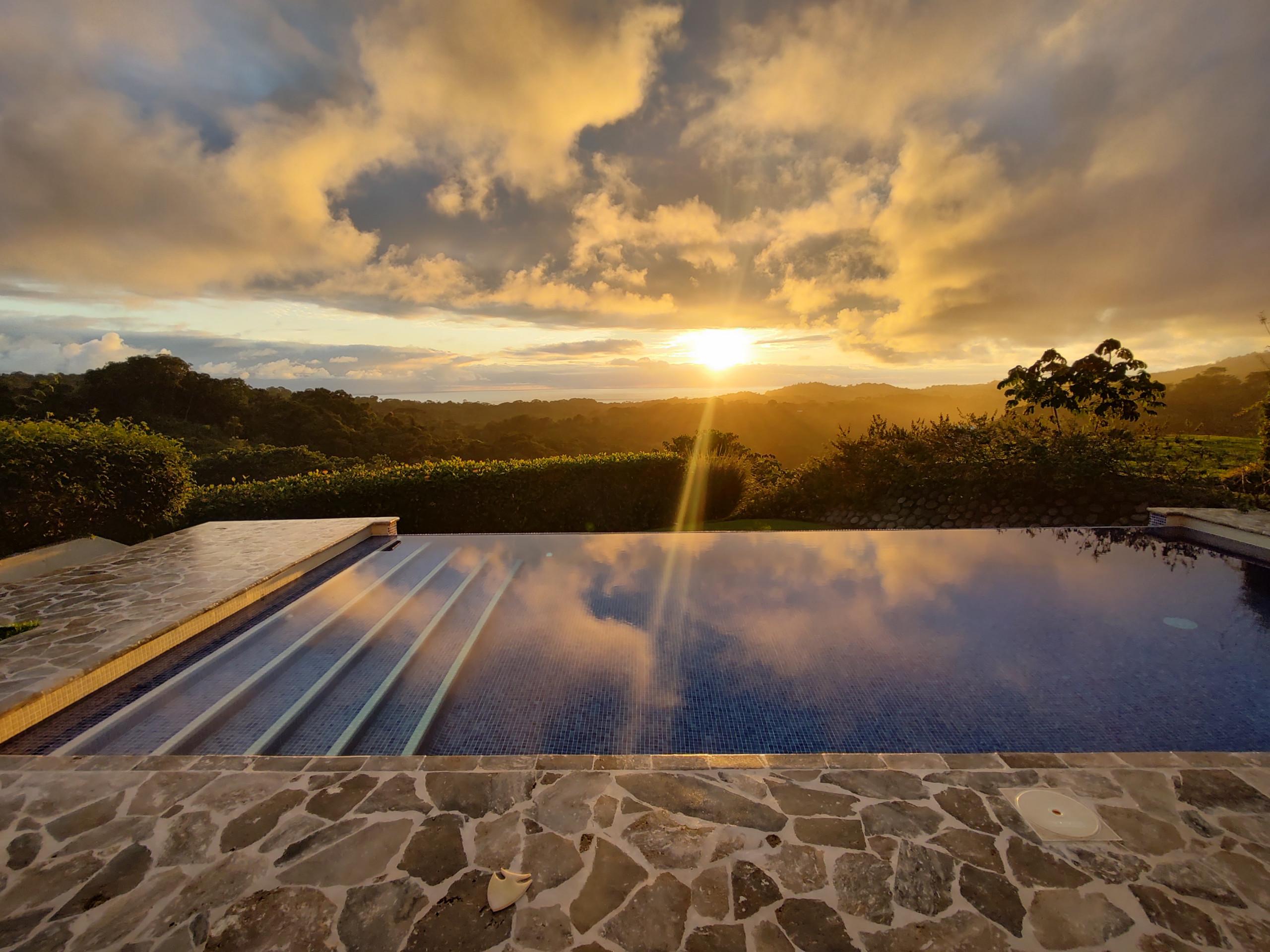 The infinity pool reflects the golden light of the sky and overlooks the expansive jungle-covered mountains extending to the distant ocean horizon.