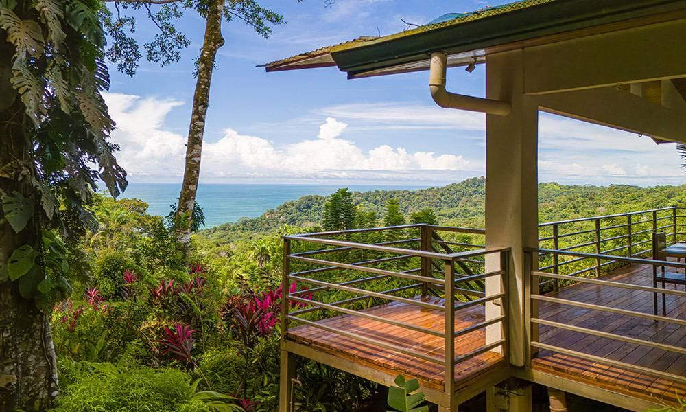 This elevated wooden deck offers an open-air living space surrounded by lush jungle, with expansive views extending to the blue ocean horizon.