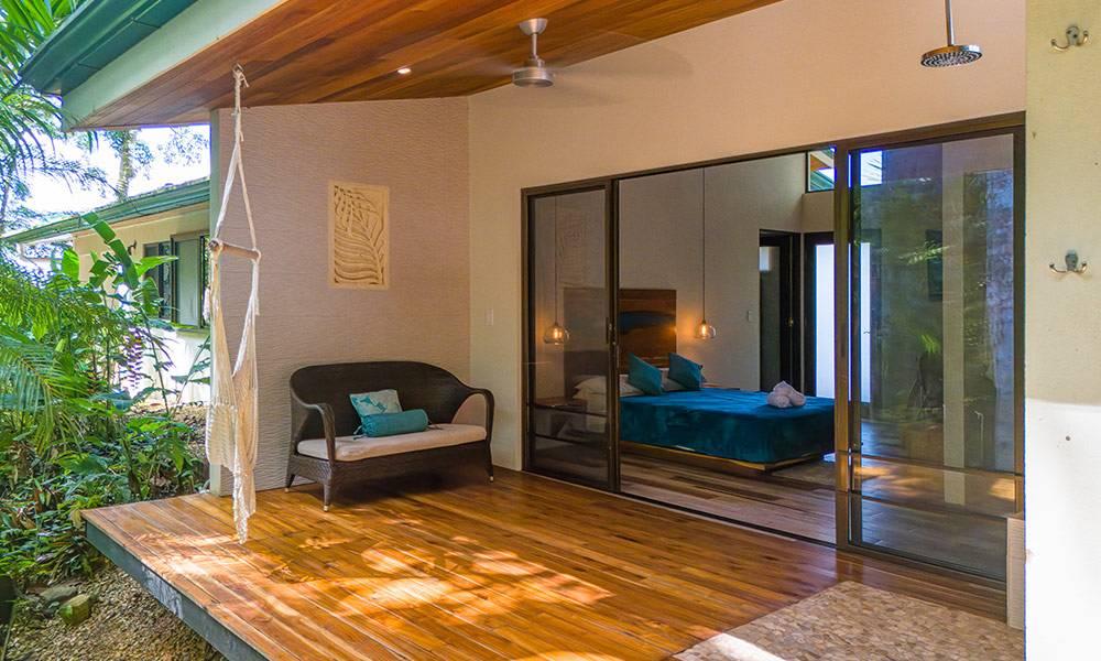 A bright bedroom suite features large glass doors opening to a private wooden terrace with jungle views and an outdoor rain shower.