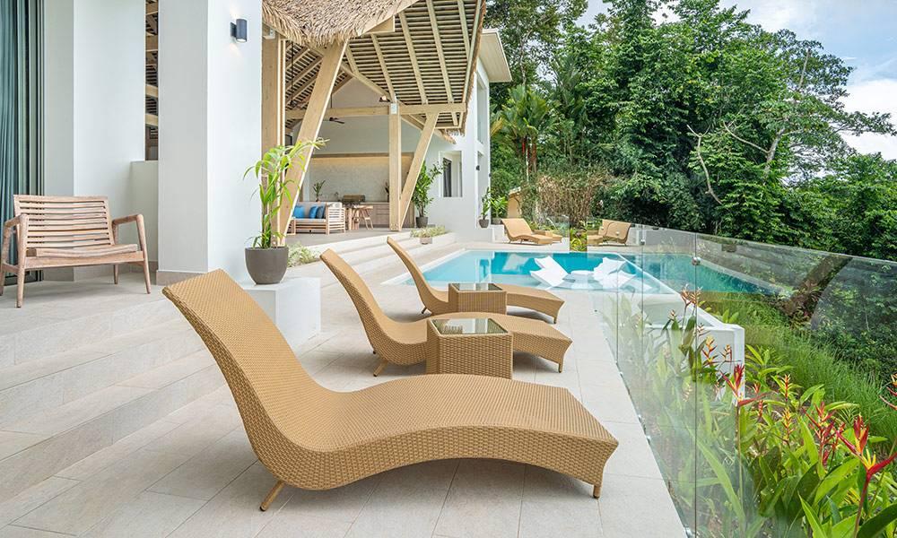 An expansive outdoor pool terrace with multiple loungers and an infinity pool offers lush jungle views from behind a clear glass railing, connecting to an open-air living area under a vaulted thatched roof.