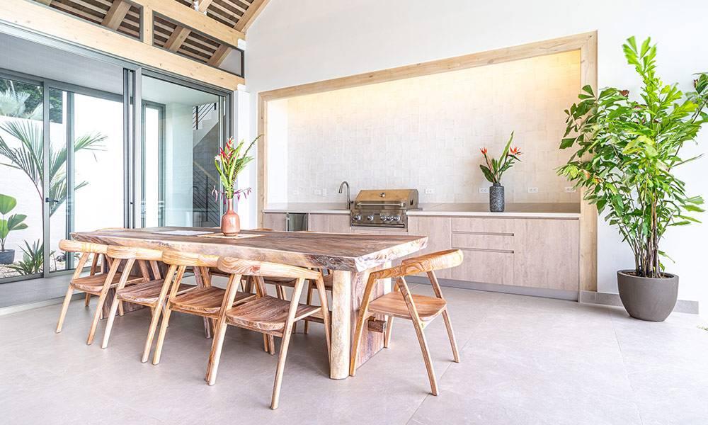 This open-air dining area features a live-edge wooden table and chairs, an integrated outdoor kitchen with a grill, and vaulted ceilings, all bathed in natural light and connected to tropical greenery through large sliding glass doors.