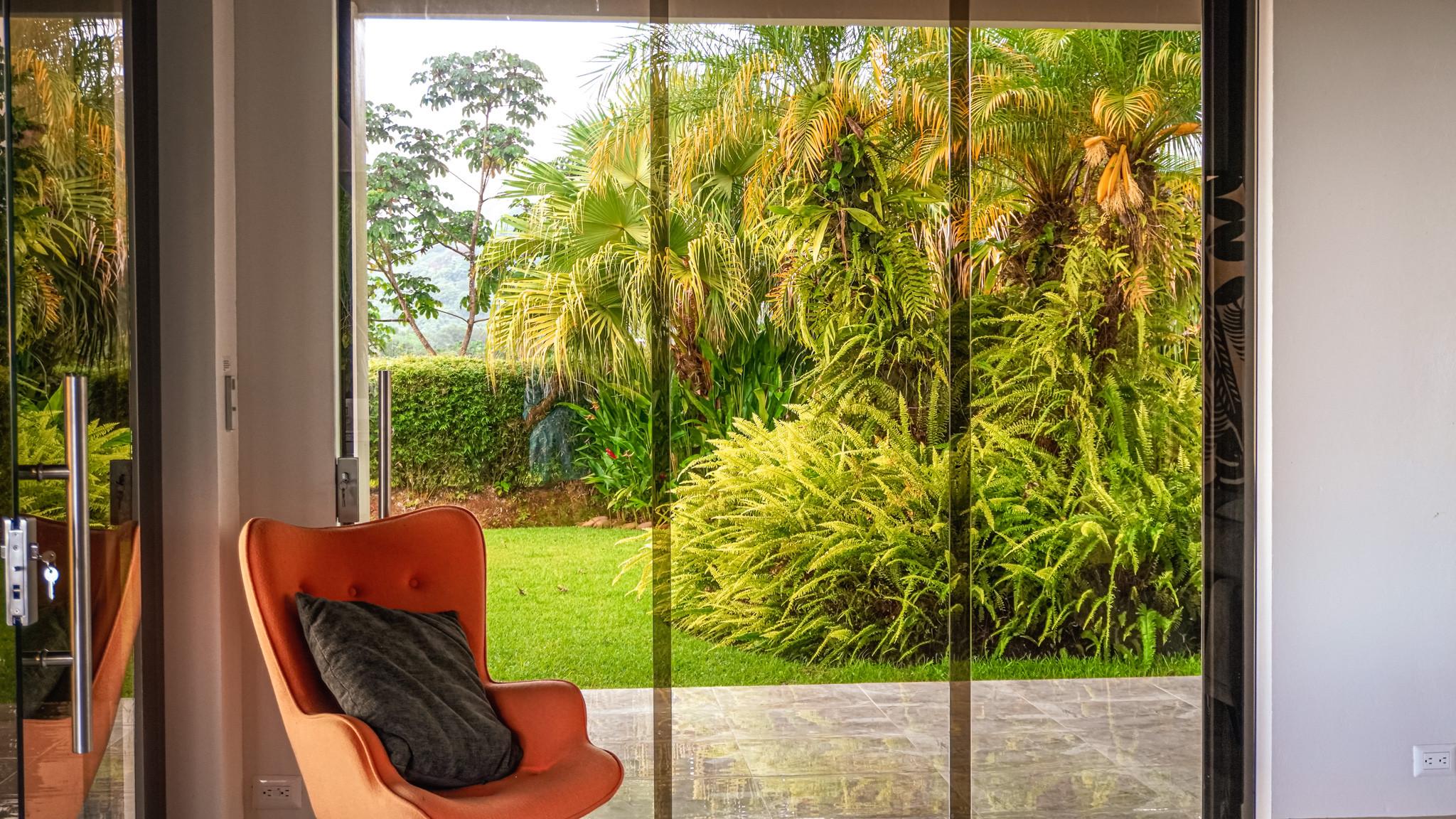 The sunlit living area opens to a lush tropical jungle garden through expansive glass doors, connecting the interior with nature.
