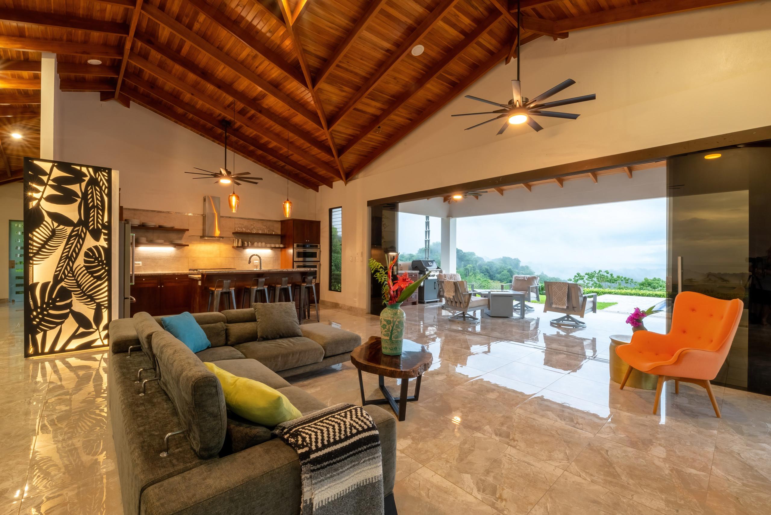 A vaulted wood ceiling crowns this open-concept living and kitchen area, opening through wide glass doors to a covered porch with views of lush jungle and distant ocean.