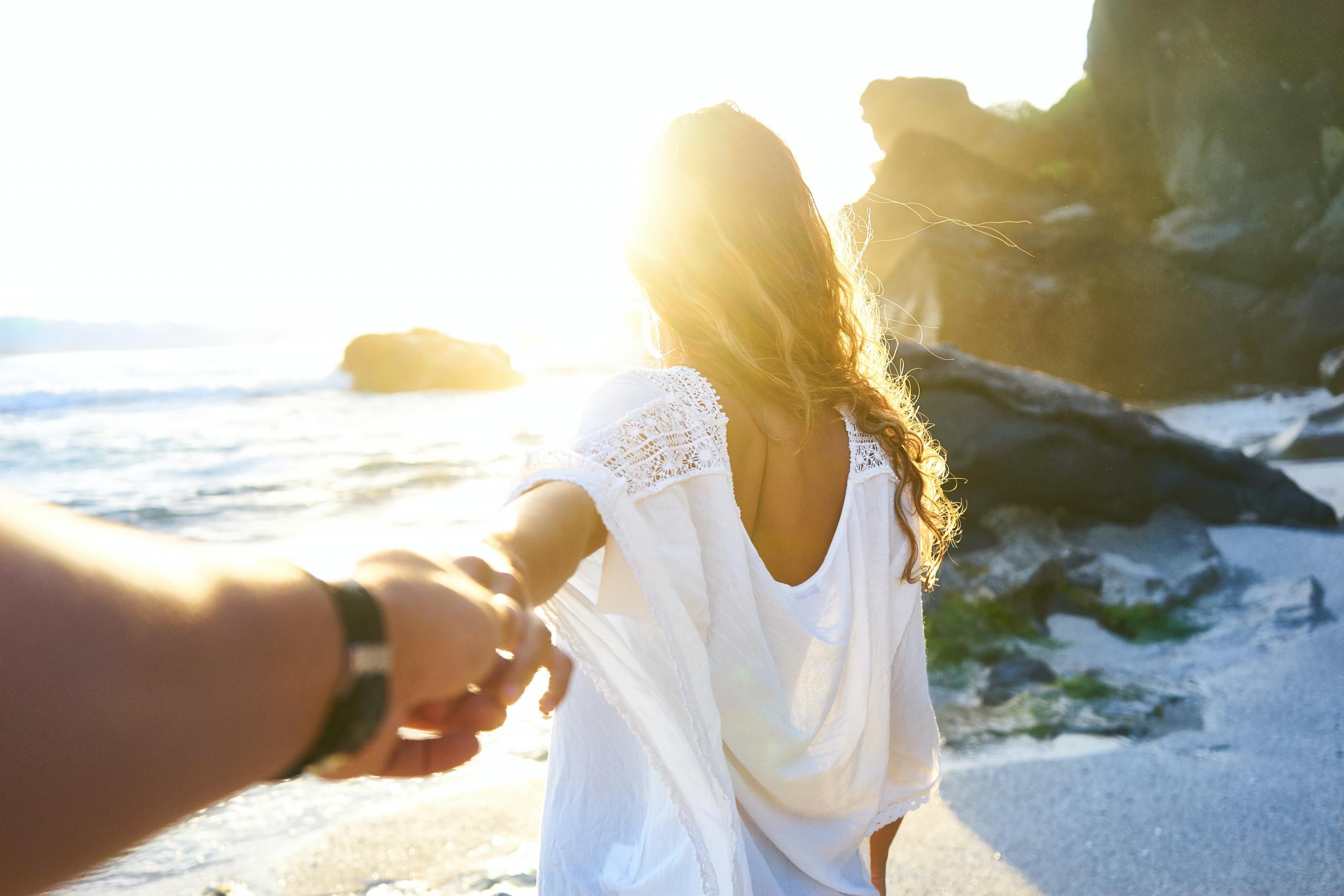 A bright sunset illuminates a person walking on a sandy and rocky beach towards the ocean, holding another person's hand.