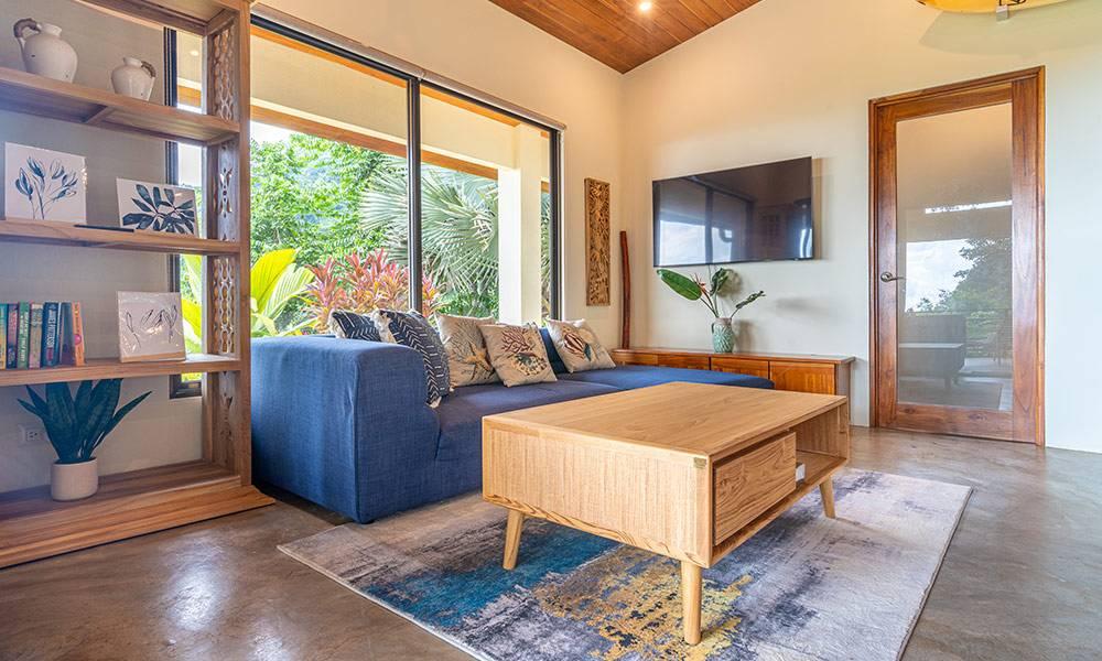 This open living area features a vaulted wood ceiling, polished concrete floor, and expansive glass doors that open to a lush jungle view, filling the space with natural light.