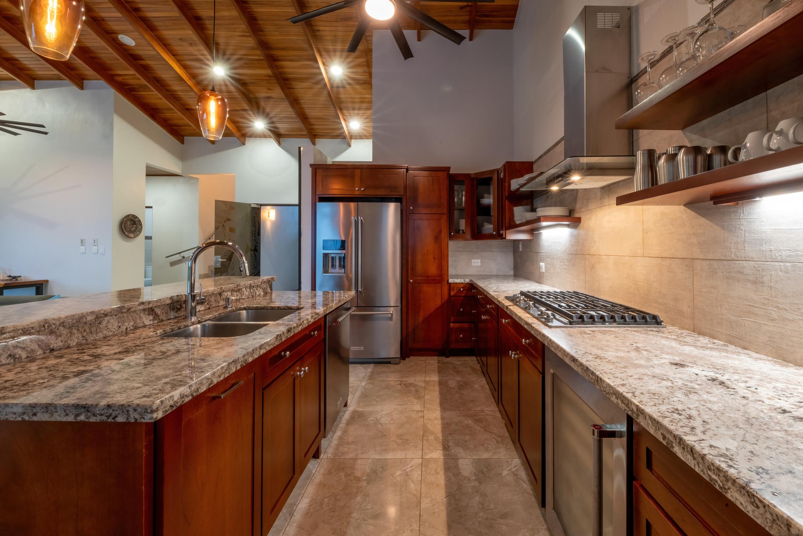 A kitchen features a large island, granite countertops, dark wood cabinetry, and stainless steel appliances under a high wood-beamed ceiling with pendant and recessed lighting, forming an open and functional space.