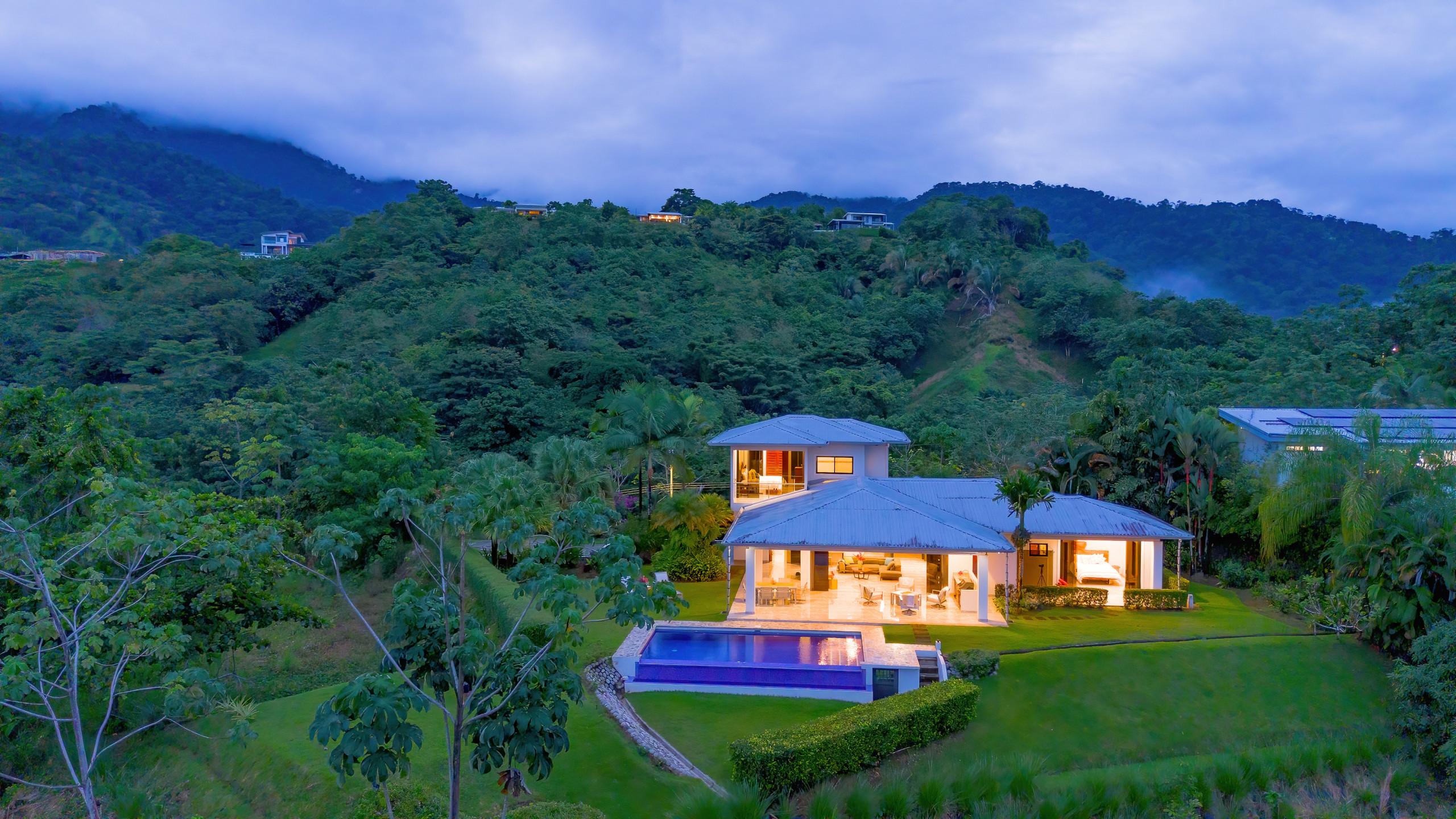 The multi-level villa is illuminated at dusk, showcasing an open-concept ground floor with a covered porch and an infinity pool, all set amidst lush, jungle-covered mountains.