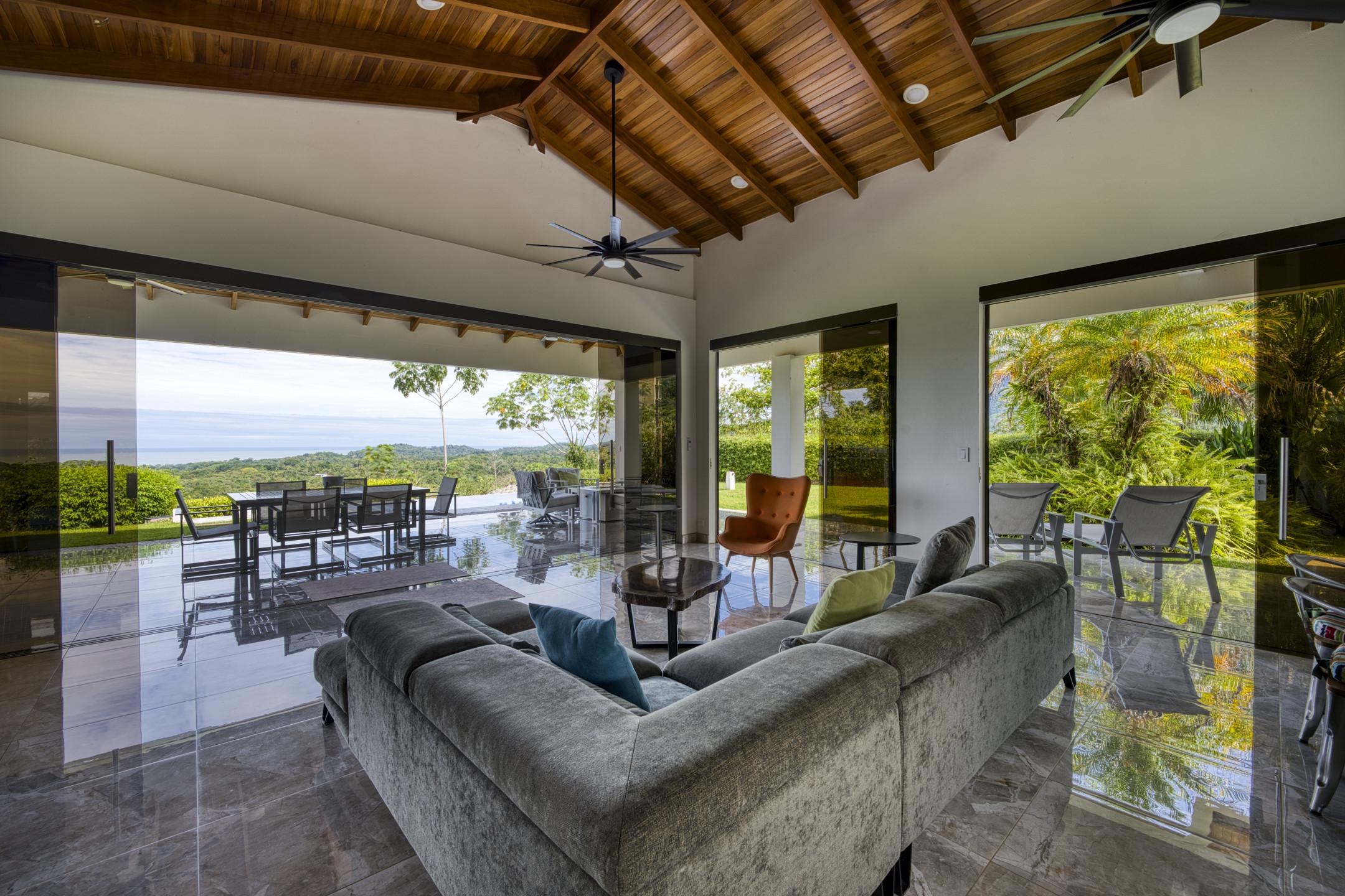 An open-concept living space with a vaulted wood ceiling and floor-to-ceiling glass walls connects seamlessly to outdoor dining, offering expansive ocean and jungle views bathed in natural light.