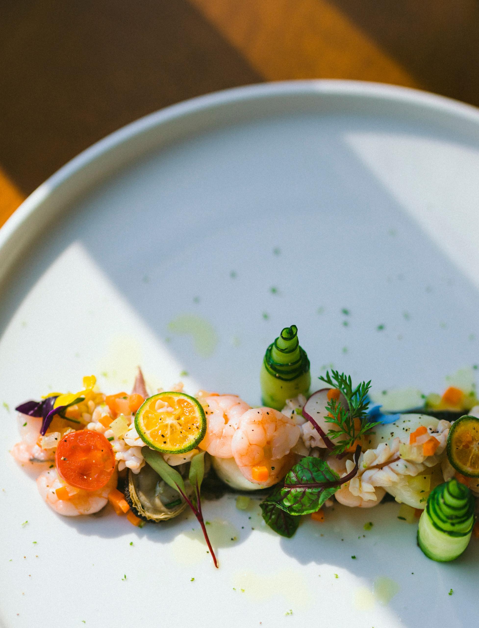A vibrant, artfully plated seafood dish, featuring shrimp, fresh vegetables, and citrus, rests on a white ceramic plate.