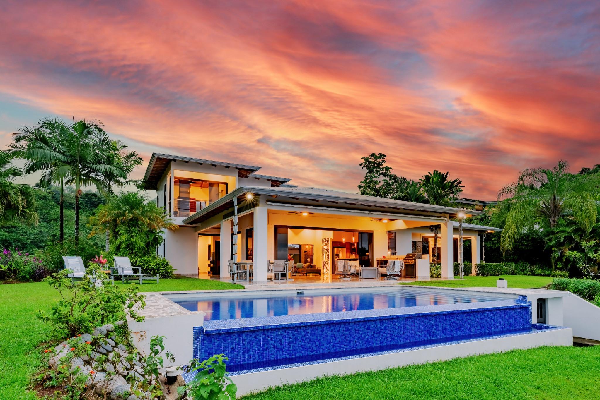 The modern multi-level villa features an illuminated open-concept ground floor connecting to a covered porch, overlooking an infinity pool and tropical jungle under a vibrant sunset sky.