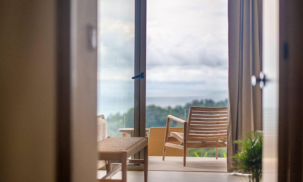 A bright bedroom opens through wide glass doors to a private terrace, revealing views of the surrounding jungle and distant ocean.
