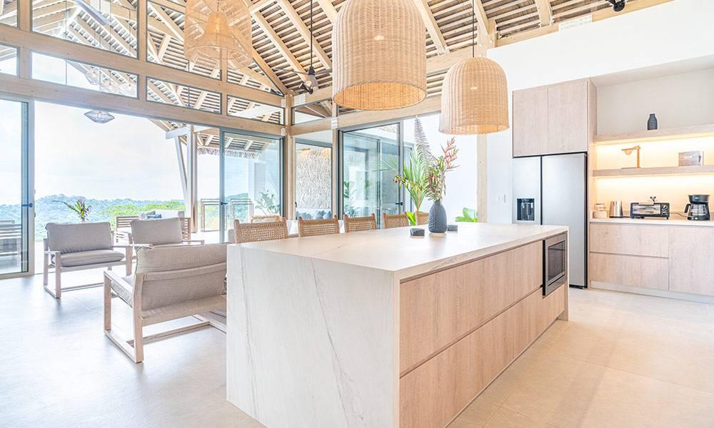 This open-plan kitchen features a large island, light wood cabinetry, and vaulted ceilings with exposed beams, receiving abundant natural light from sliding glass doors that offer distant jungle views.