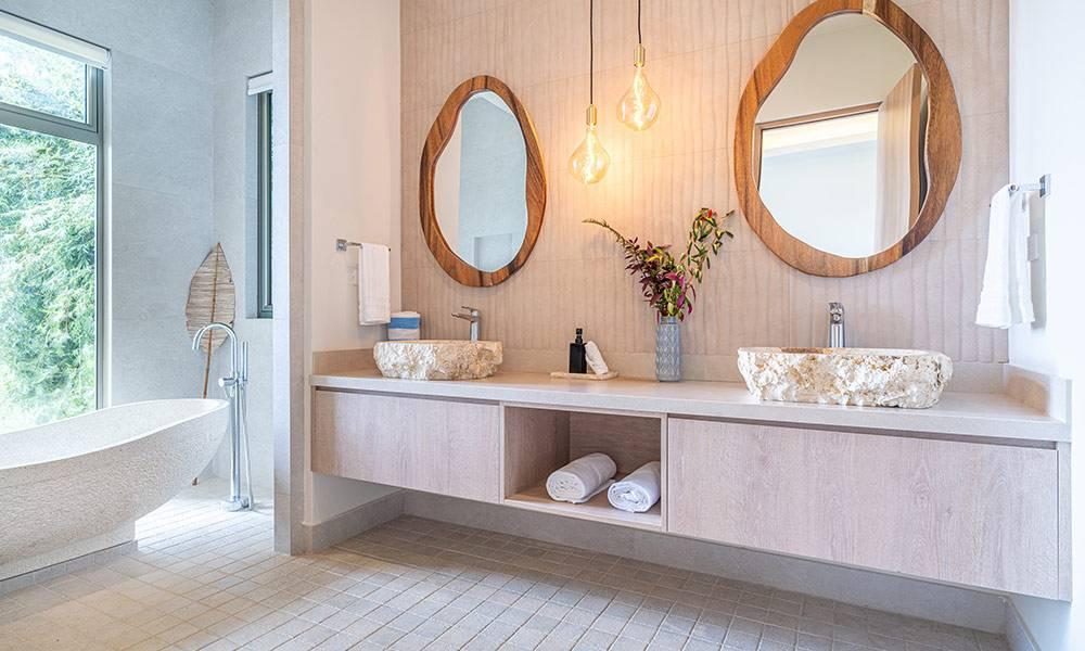 A bright bathroom features a double vanity with textured stone vessel sinks, natural wood-framed mirrors, a freestanding bathtub, and a large window providing natural light and jungle views.