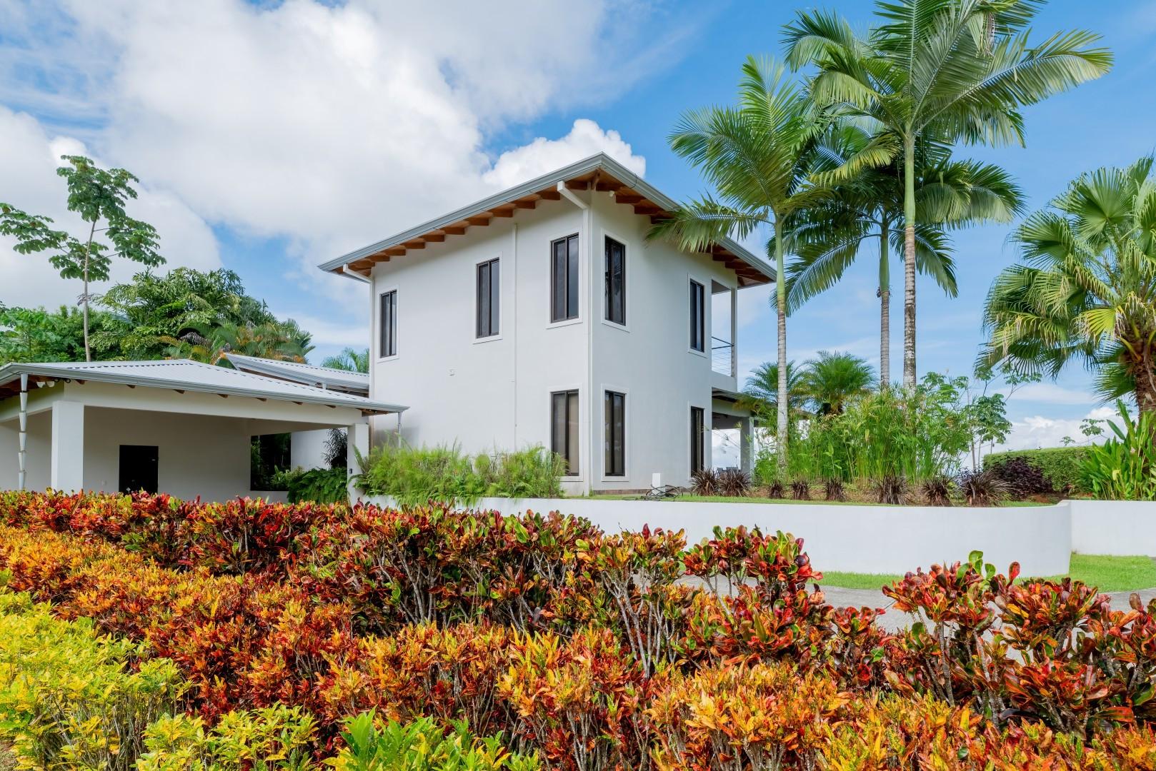 A modern, two-story white villa with dark-framed windows, a covered parking area, and an upper-level balcony is nestled within vibrant tropical landscaping and palm trees under a bright blue sky.