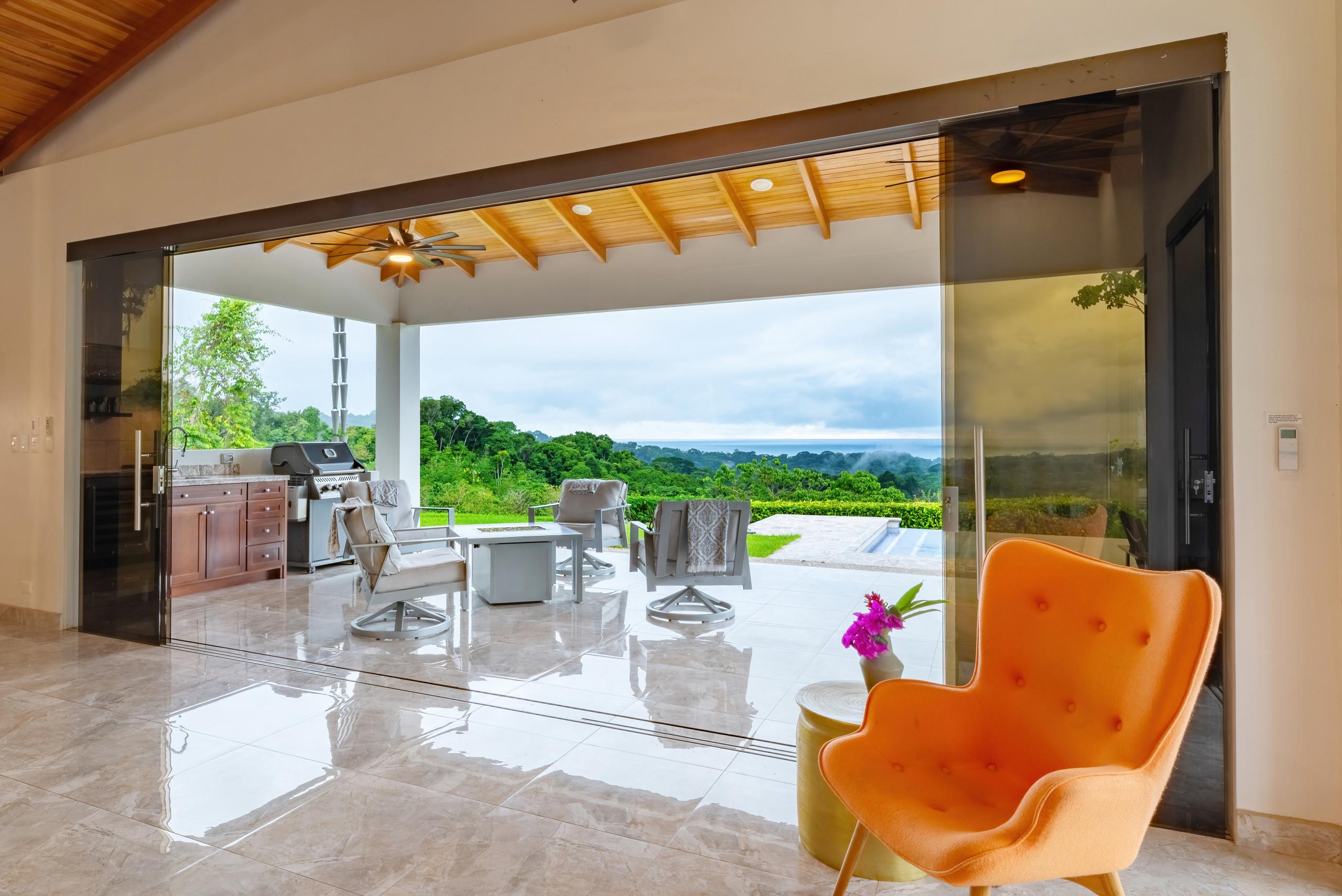 Wide sliding glass doors connect the interior to a covered terrace featuring a slatted wood ceiling, outdoor lounge seating, and a barbecue area, all overlooking lush jungle and the distant ocean.