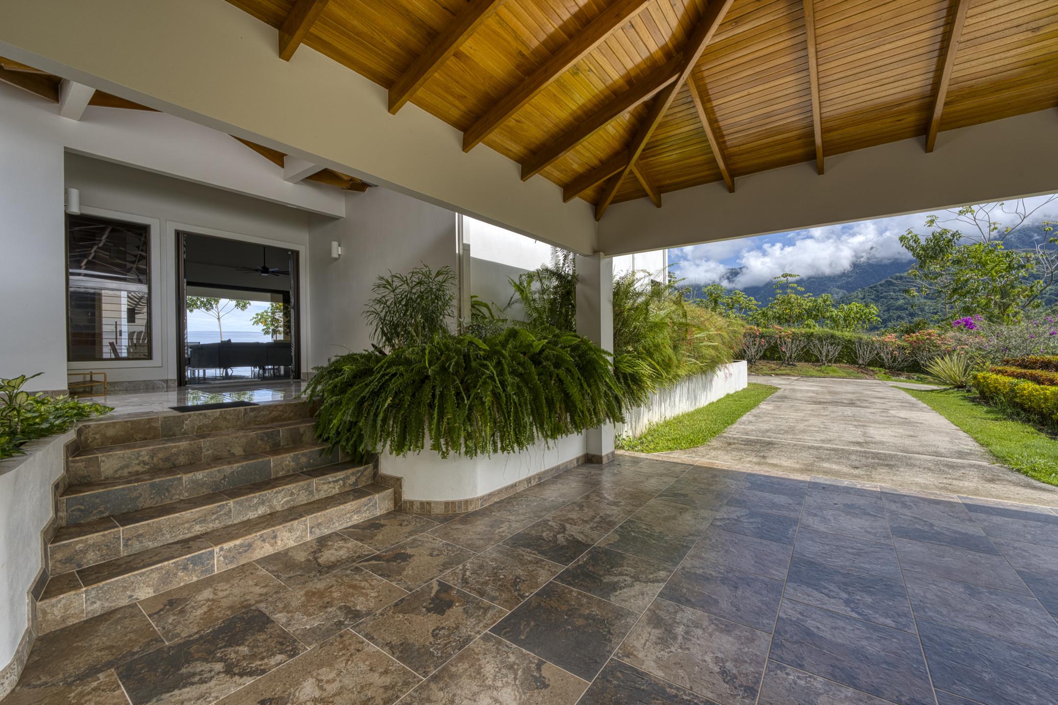 This covered villa entrance features a natural wood plank ceiling, tiled steps, and lush tropical plants, with a clear view of the ocean and mountains through the glass doors.