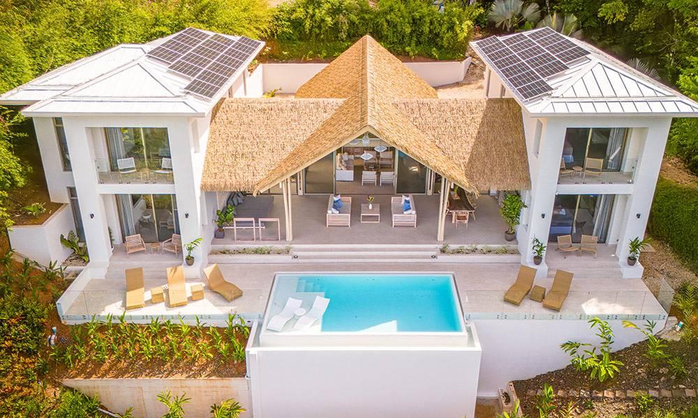 This modern villa features white structures, a central open-air thatched living area, and an infinity pool on a spacious terrace, all set within the lush tropical jungle.